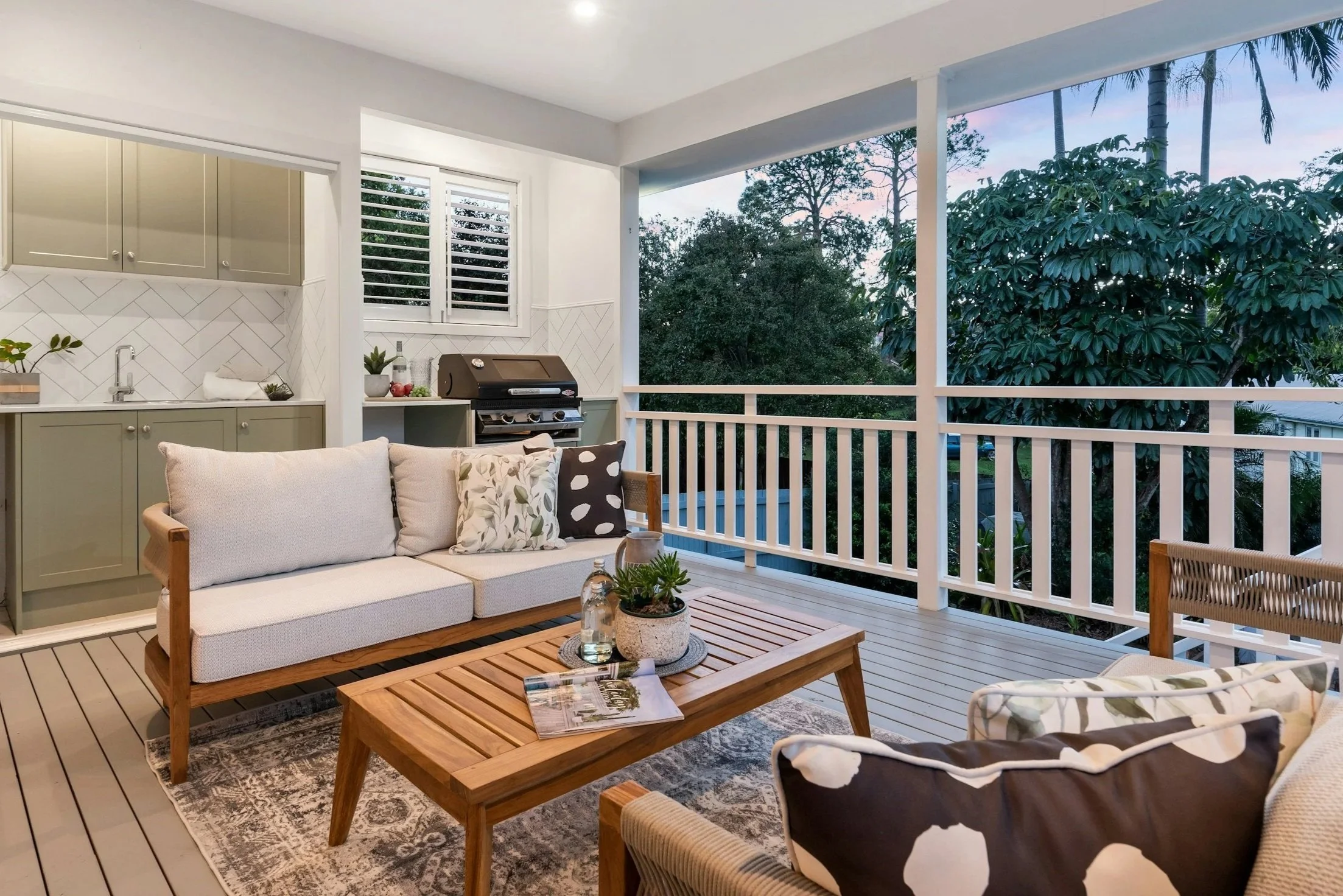 A covered balcony with white railing and wooden floor, furnished with a beige sofa, patterned pillows, a wooden coffee table with a plant and magazines, a wicker chair, green outdoor cabinets, a grill, and a window overlooking trees and a sunset sky.