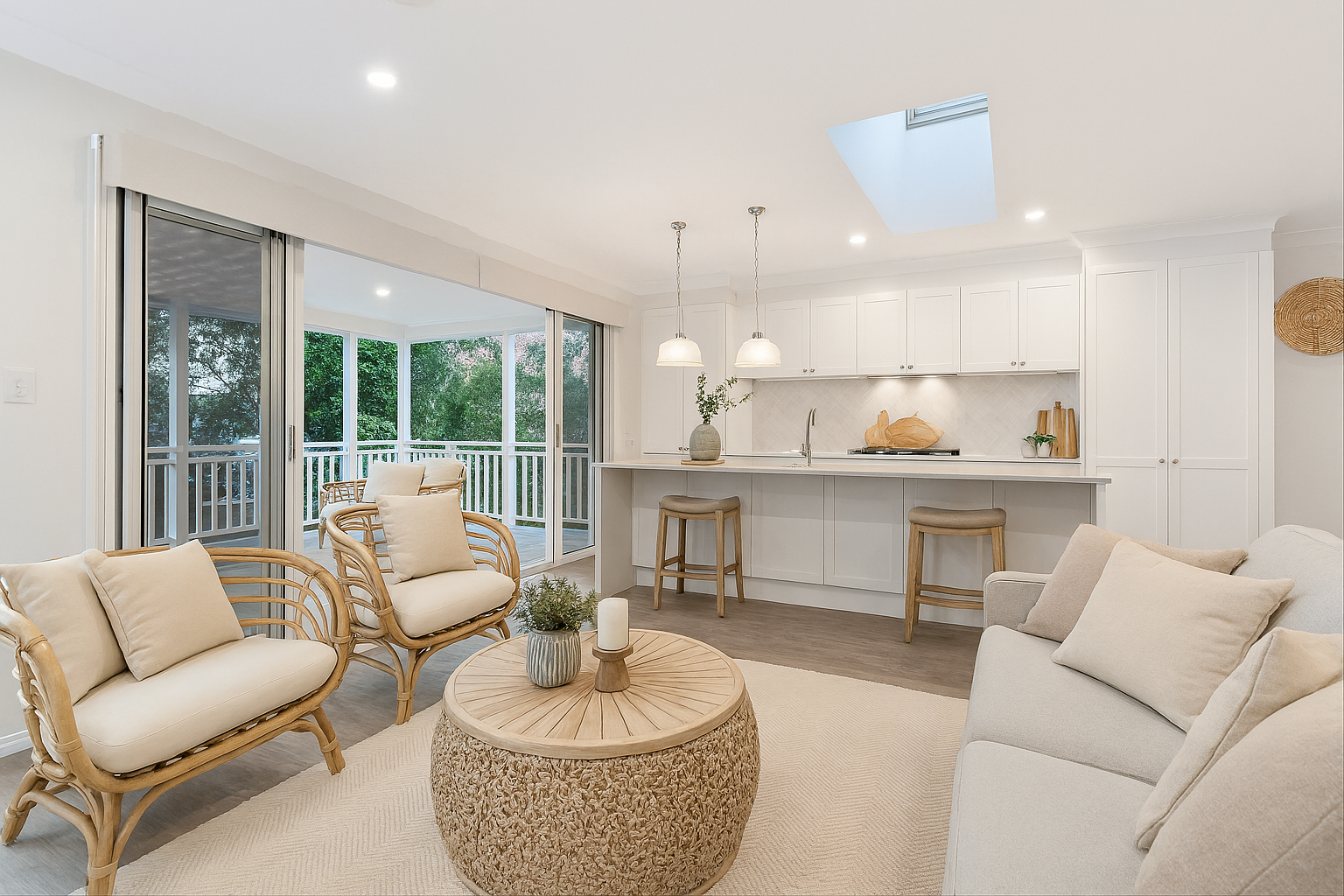 Townsville rental property. Living room with natural light, a beige sofa, a round textured coffee table with a plant, a white kitchen with a breakfast bar, bar stools, pendant lights, and a balcony with outdoor seating and greenery visible outside.