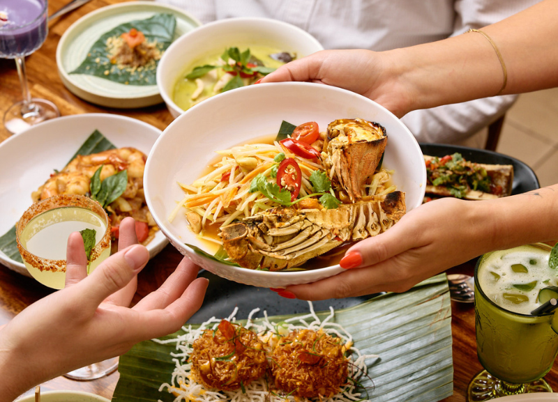 photo of a person's hands passing a dish to someone else over a table with several thai dishes