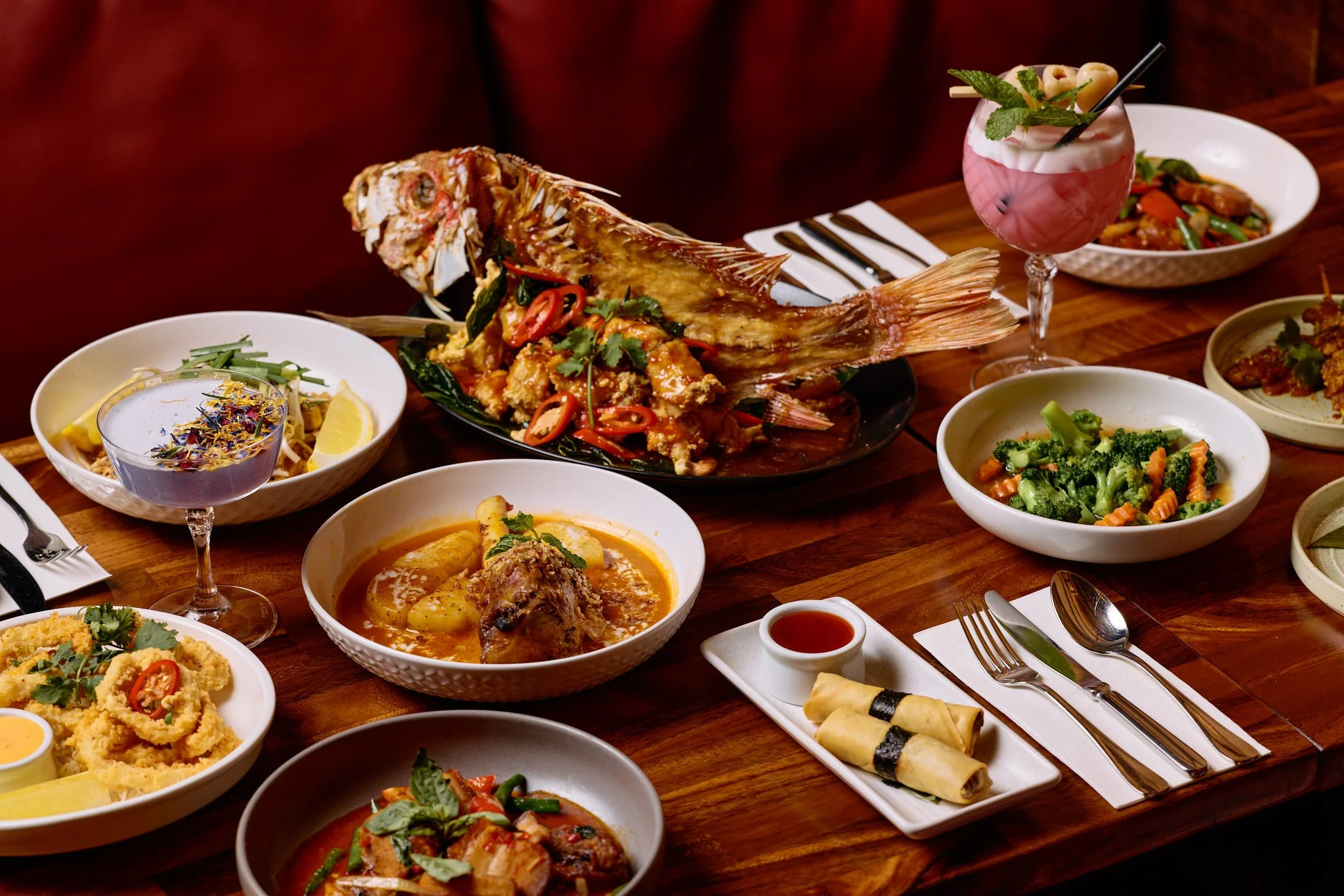 Phot of a table with several Thai dishes on it. From whole cooked fish to spring rolls, massaman curries and a cocktail.