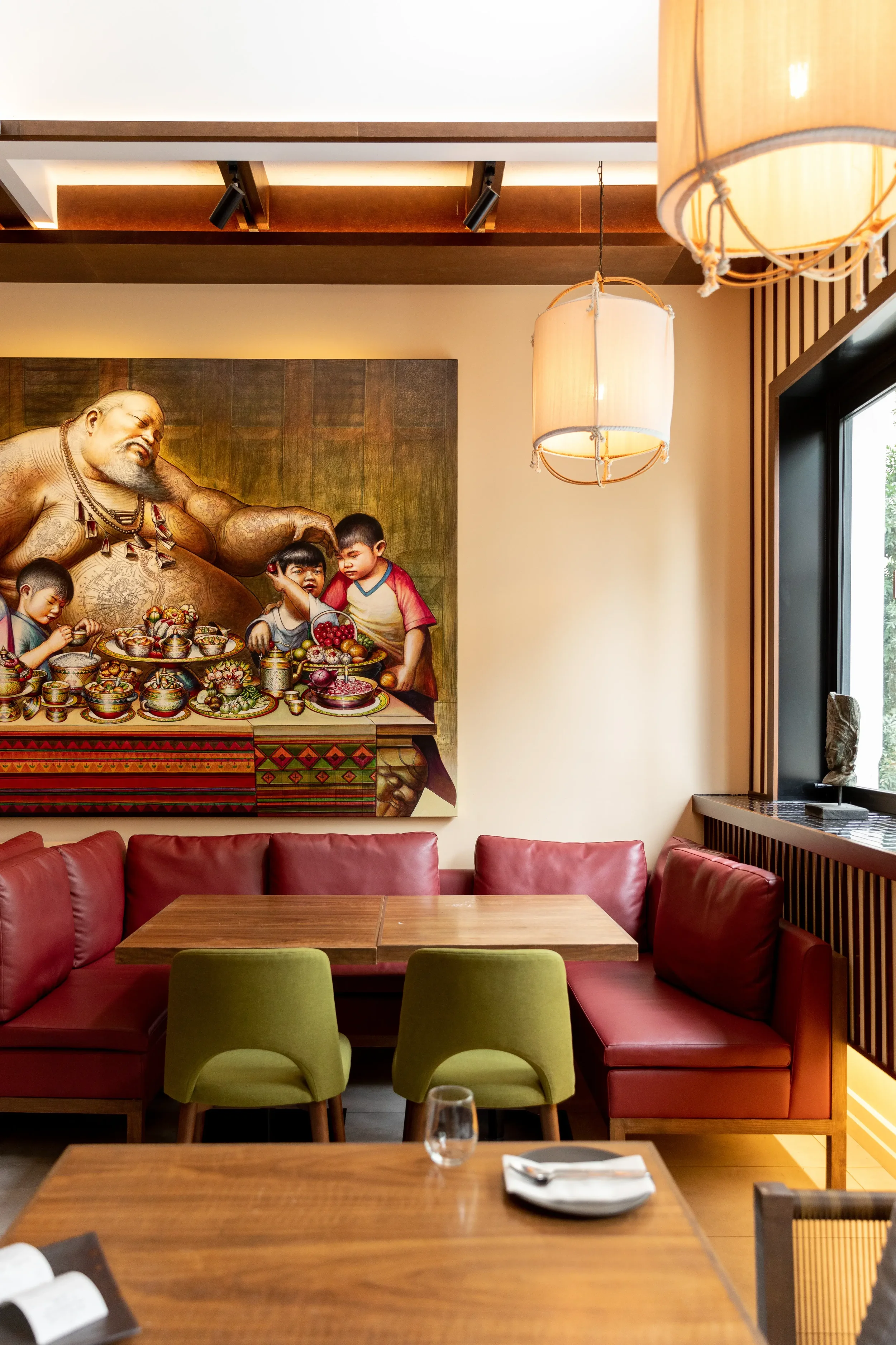 Interior view of a restaurant with wooden tables, red and green chairs, a large window, two hanging lamps, and a large painting on the wall depicting a traditional scene with a man and children preparing food.