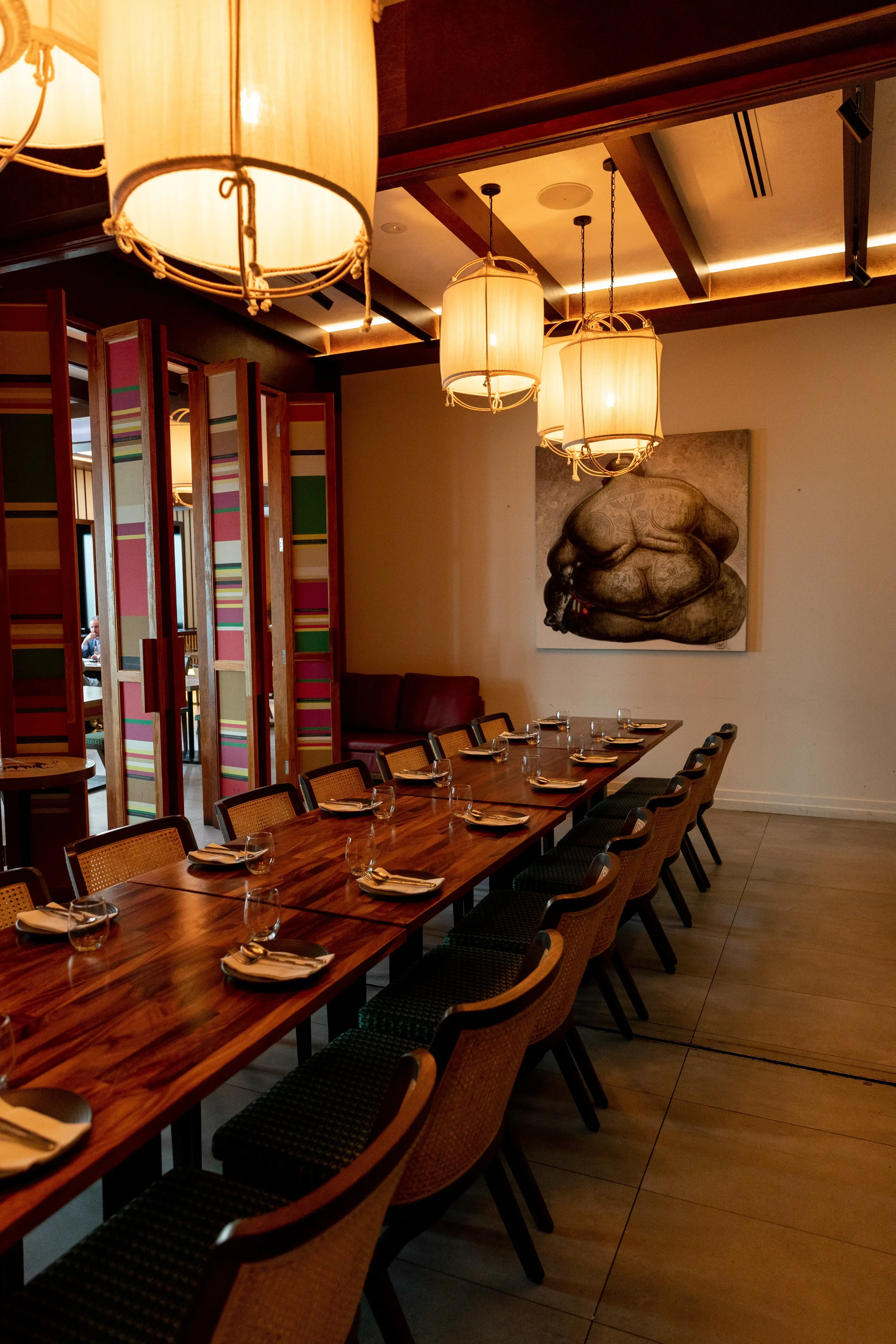 A dining room with a long wooden table set with plates, glasses, and napkins, surrounded by chairs. There are hanging pendant lights and a large abstract black-and-white artwork on the wall.