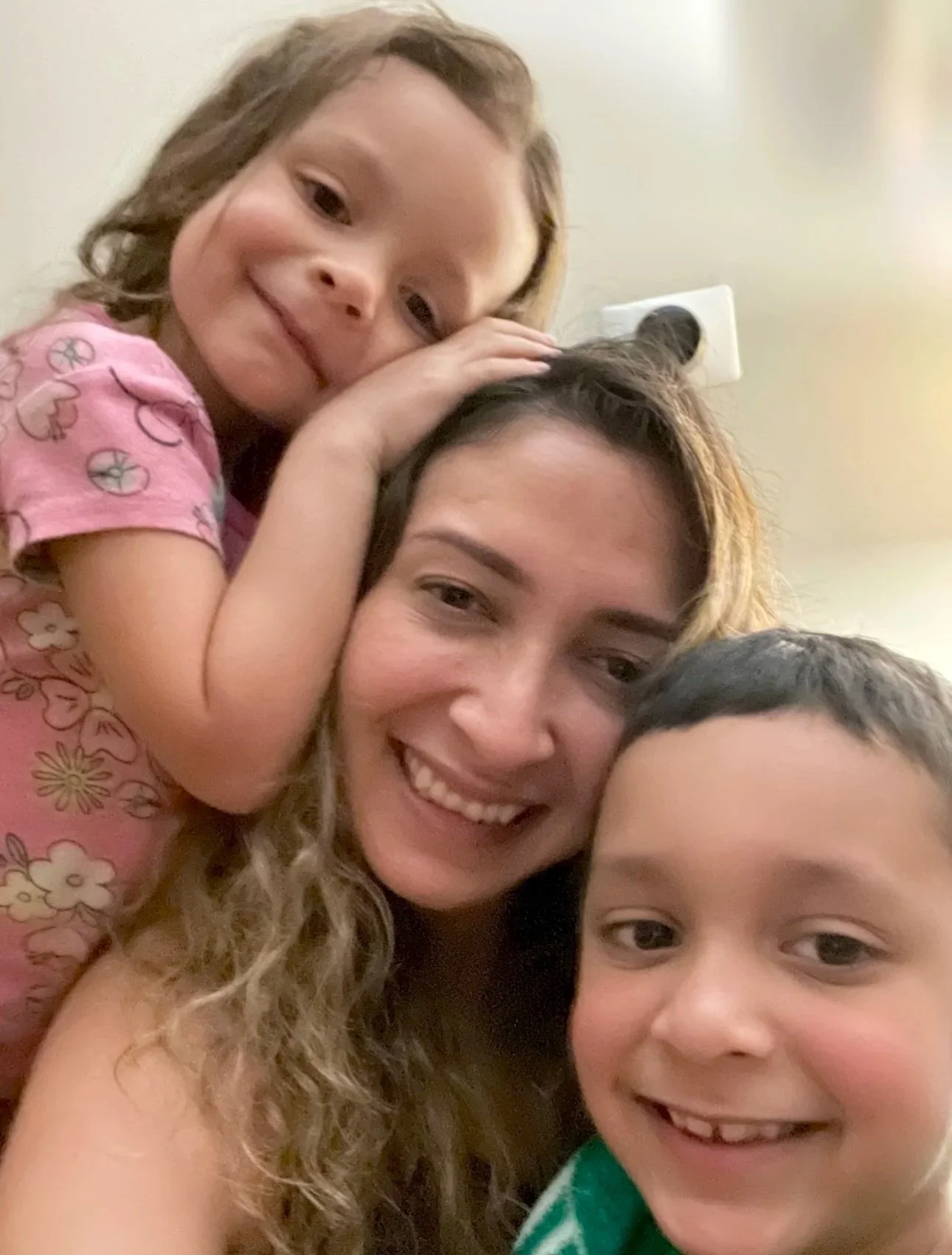Smiling woman with two children, a girl on top and a boy in front, taking a selfie indoors.