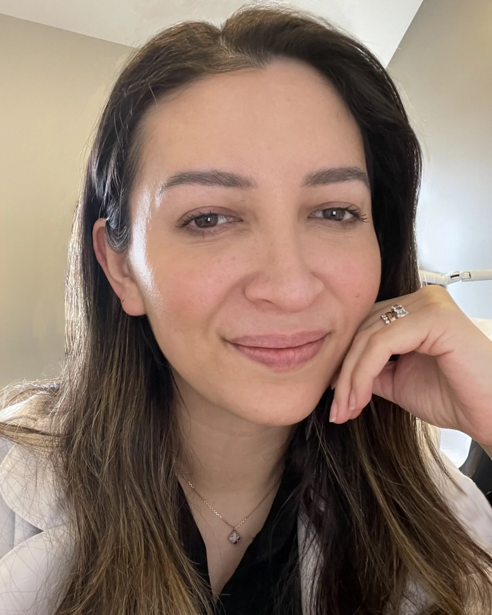 Close-up of a woman with dark hair, smiling, resting her chin on her hand, wearing jewelry, indoors.