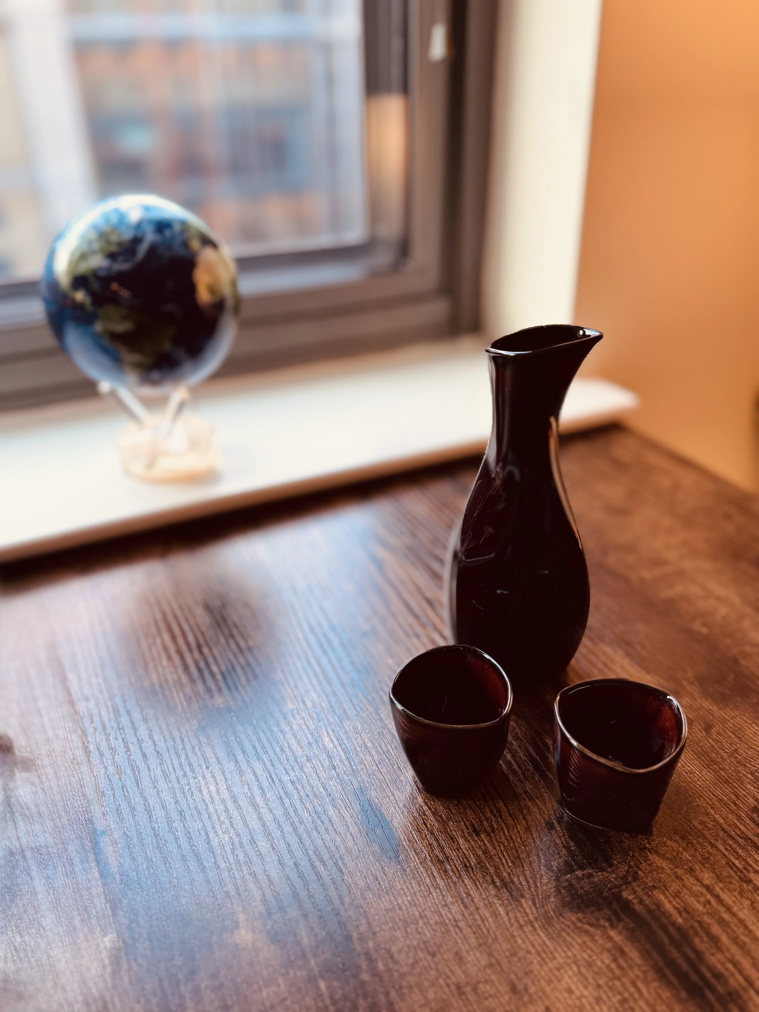 Black ceramic sake set with a sake bottle and two cups on a wooden table, with a window in the background showing a blurred view of buildings and a globe on the windowsill.