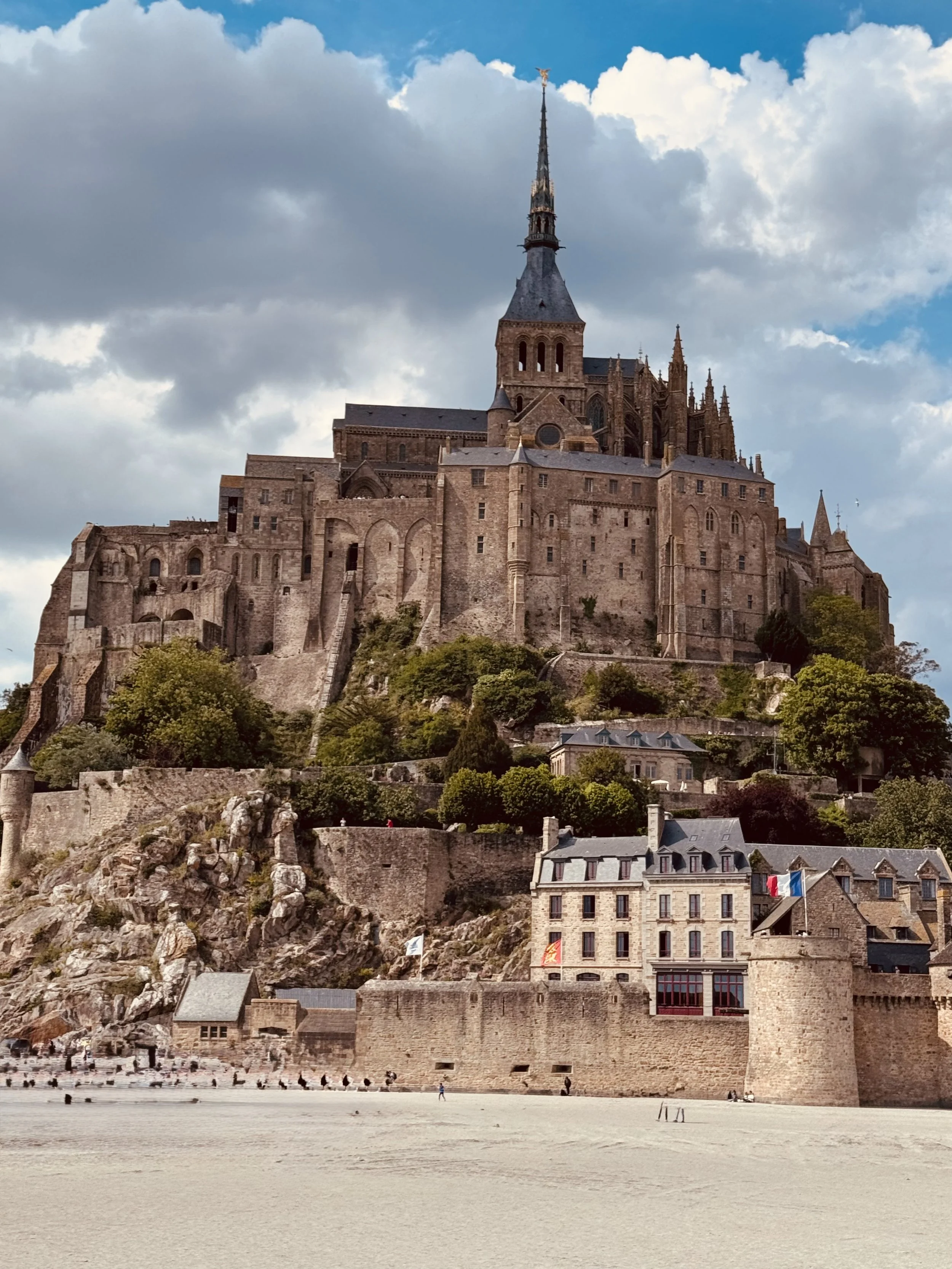 A large medieval castle atop a hill, with a beach and historical buildings at the base, under partly cloudy skies.