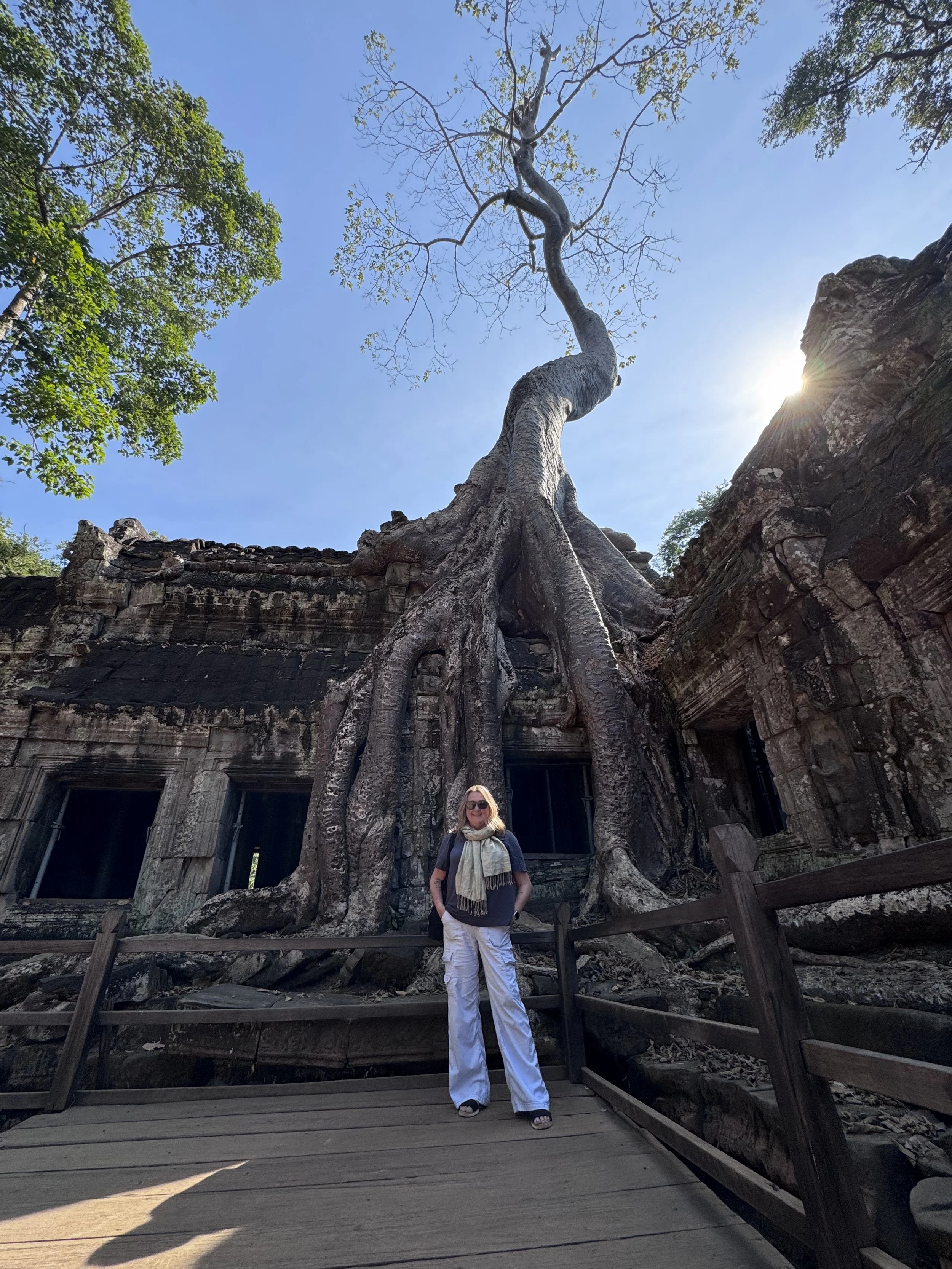 A woman standing on a wooden platform in front of an ancient temple entangled by large tree roots, with green trees and a blue sky in the background, and sunlight peeking from behind the temple.