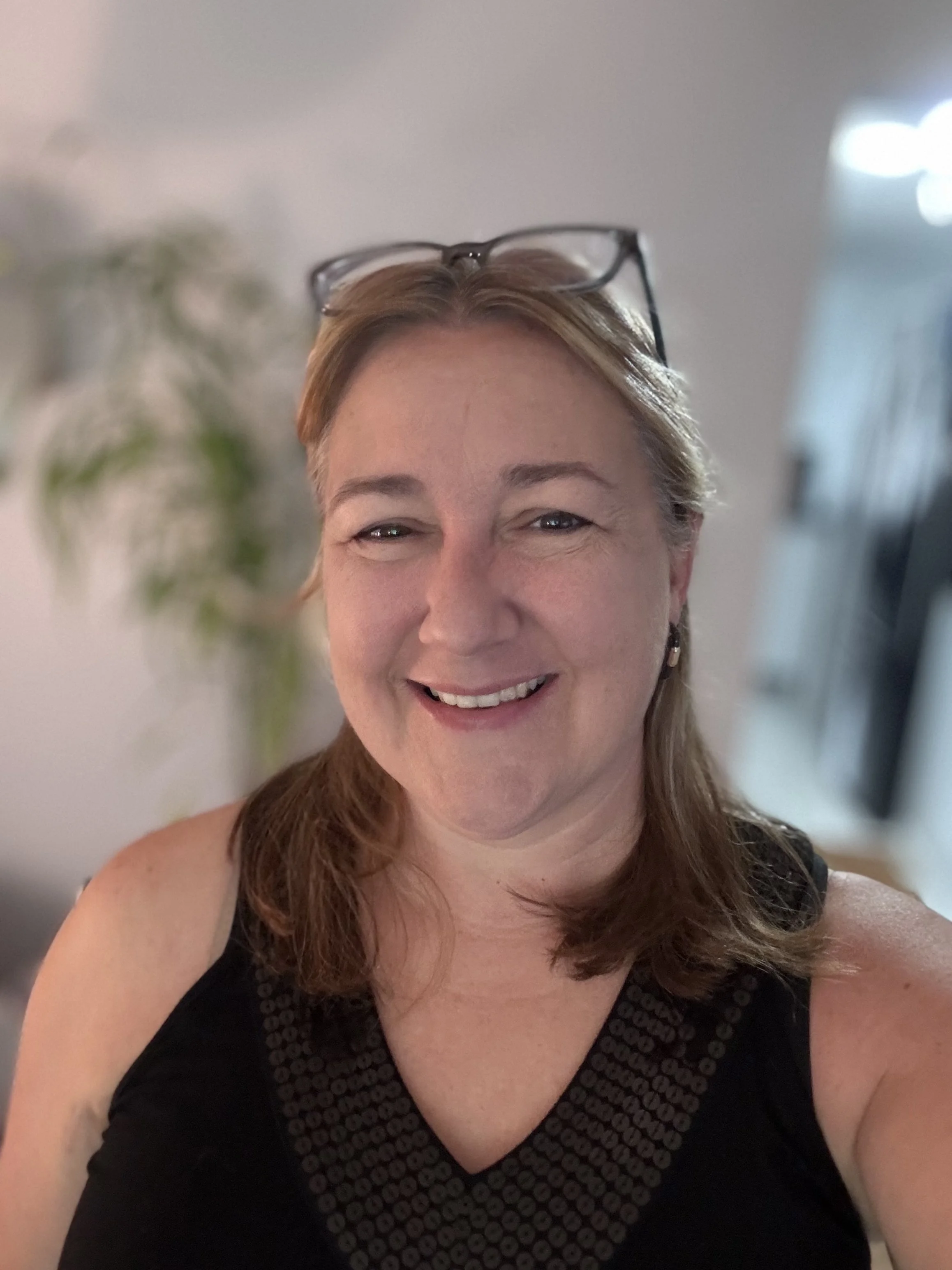 A woman with glasses on her head, smiling at the camera, wearing a black sleeveless top, standing indoors with a blurred background of a plant and a door.