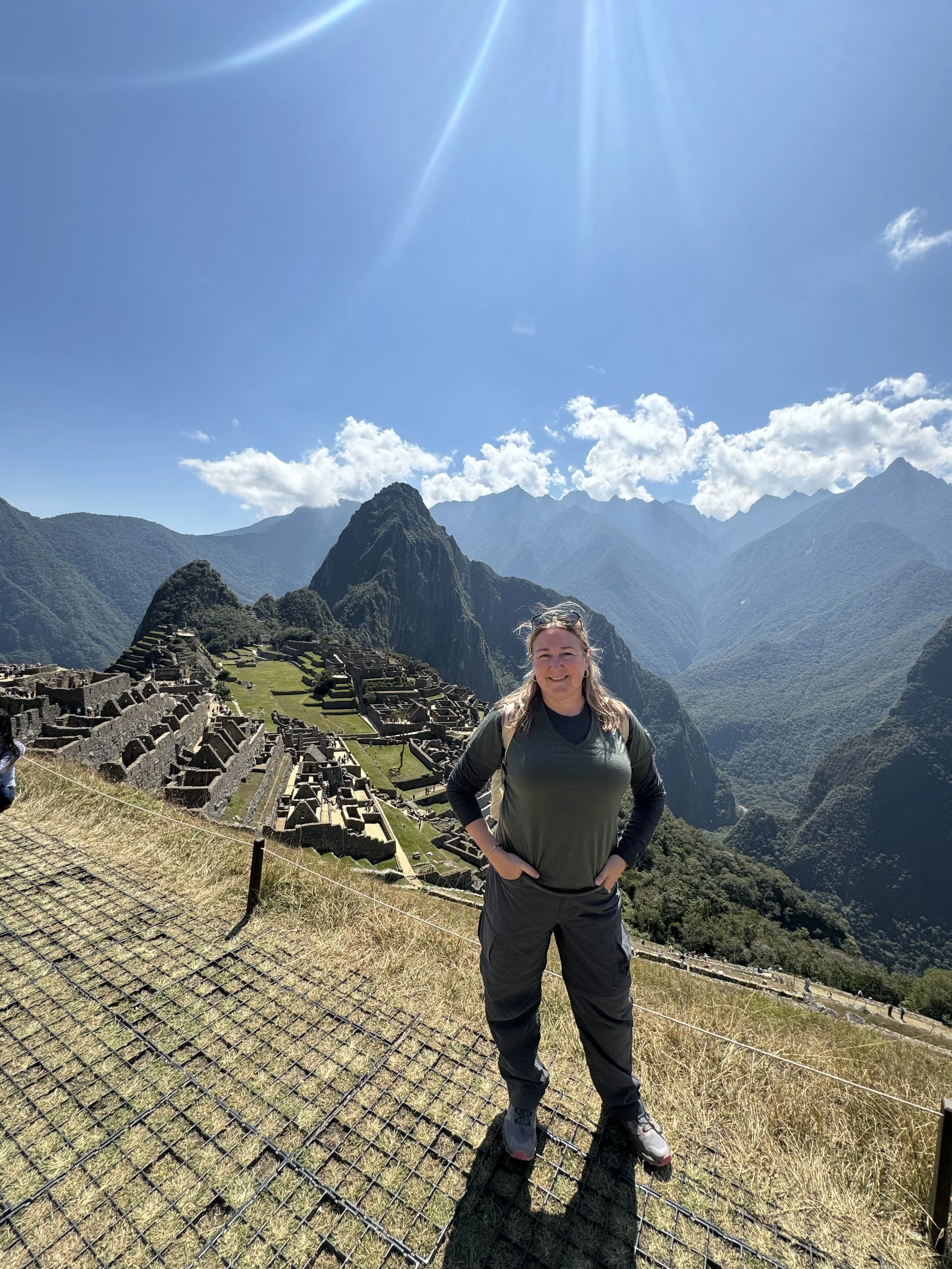 A woman standing on a grassy hill with Machu Picchu ruins and mountains in the background under a sunny sky.
