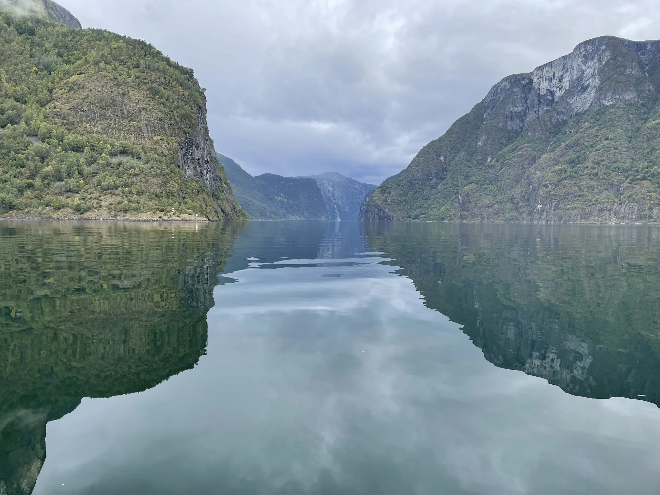 A calm fjord with towering mountains on both sides, with reflections in the water and cloudy sky overhead.