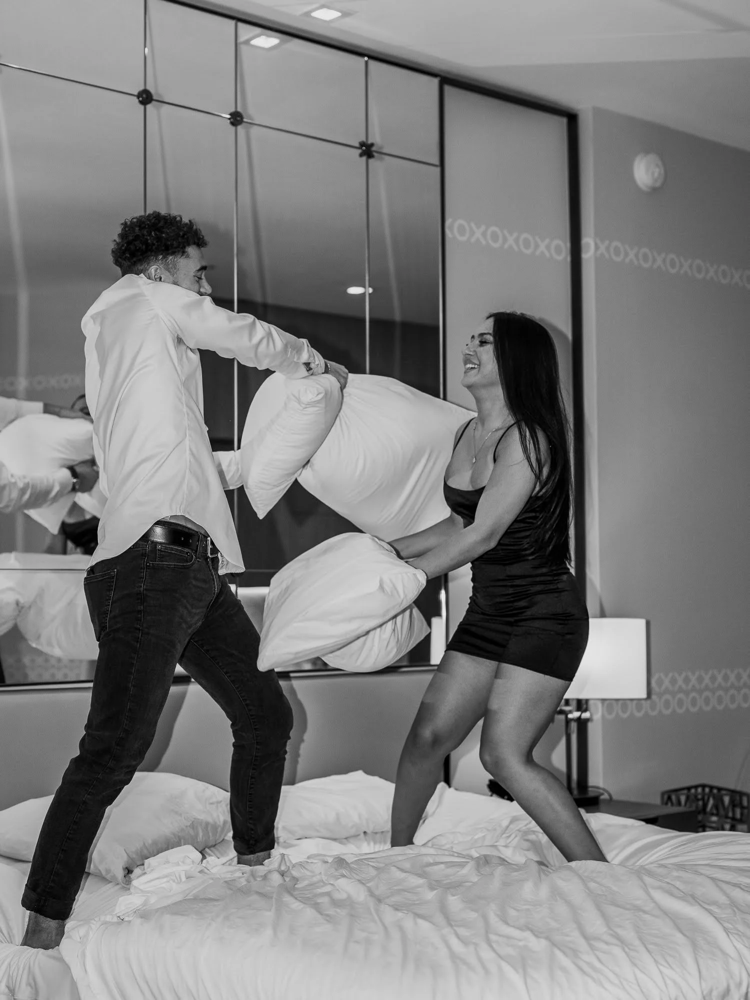 A man and woman having a pillow fight in a hotel room, both smiling and standing on a bed with pillows, with a mirror and a lamp in the background.