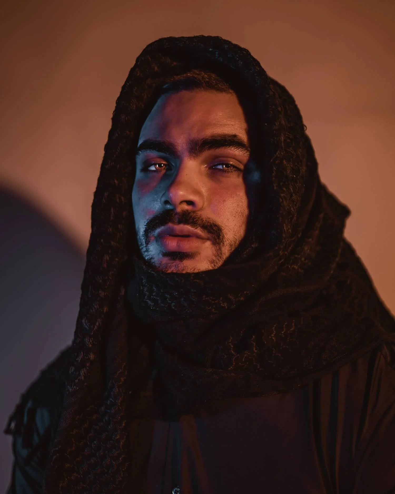 A man with a dark scarf wrapped around his head and neck against a warm, gradient background.
