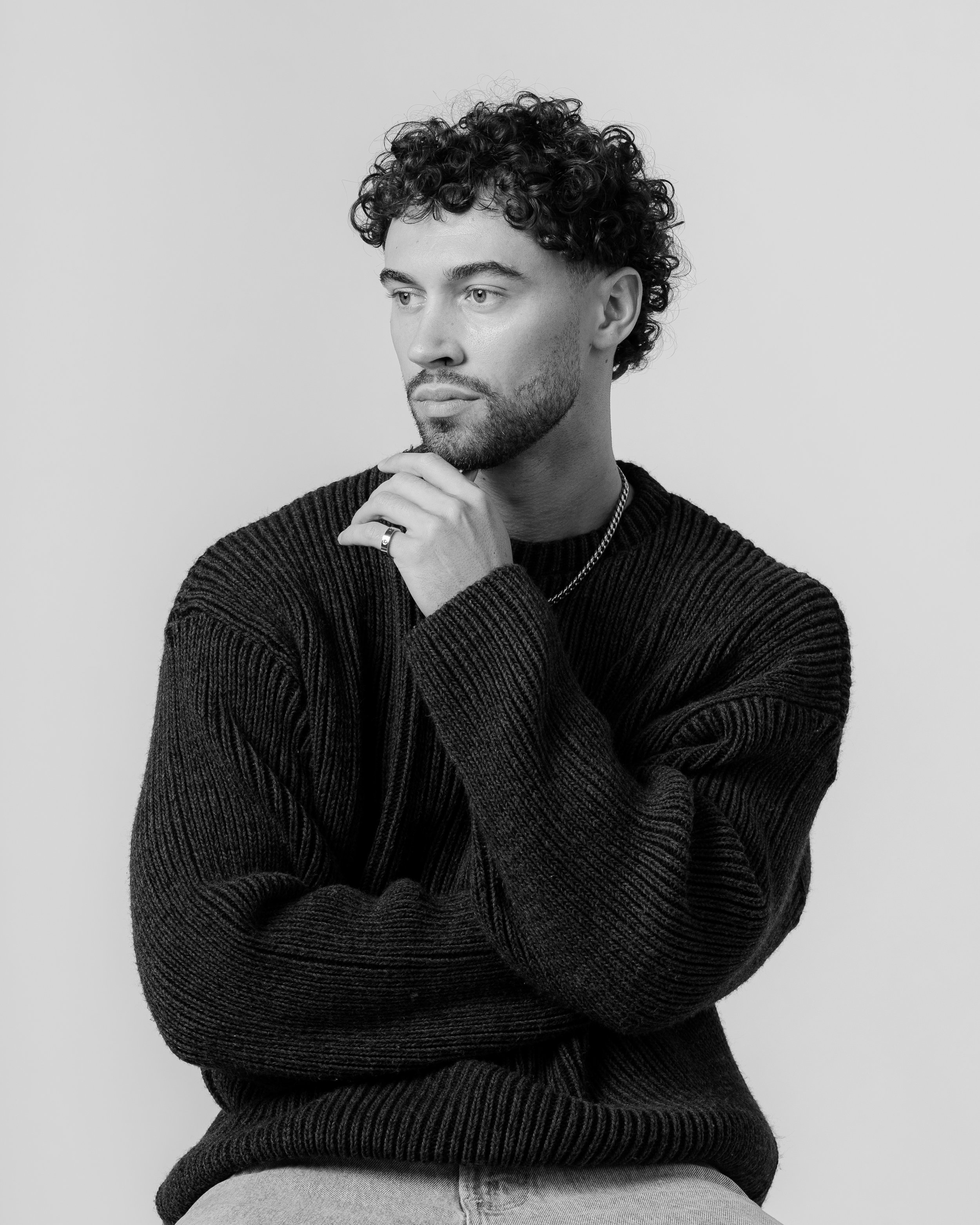 Black and white portrait of a young man with curly hair and a beard, wearing a dark sweater, with one hand touching his chin and looking thoughtfully to the side.
