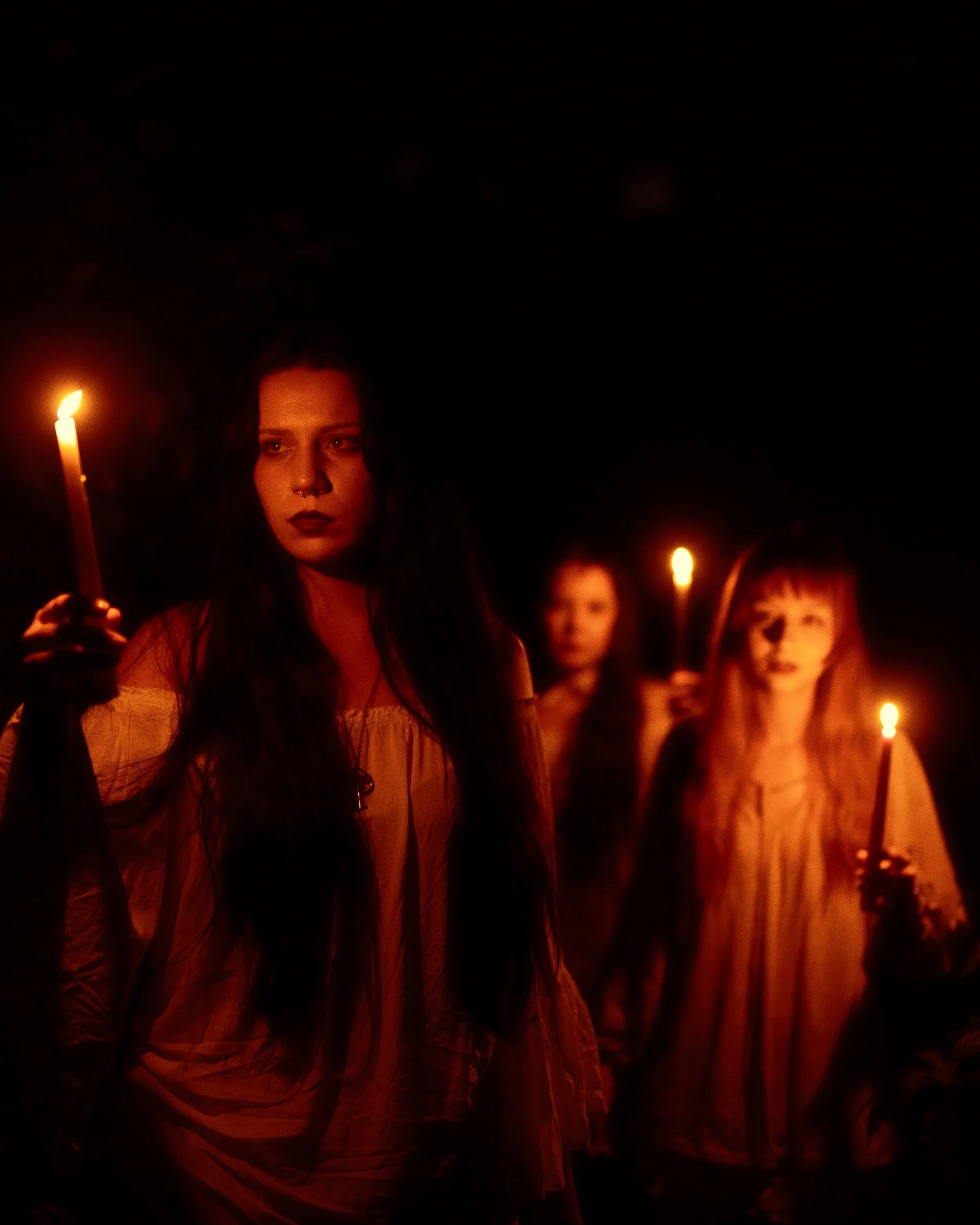 Three women with long hair, holding lit candles, walking in darkness, wearing loose, light-colored clothing.