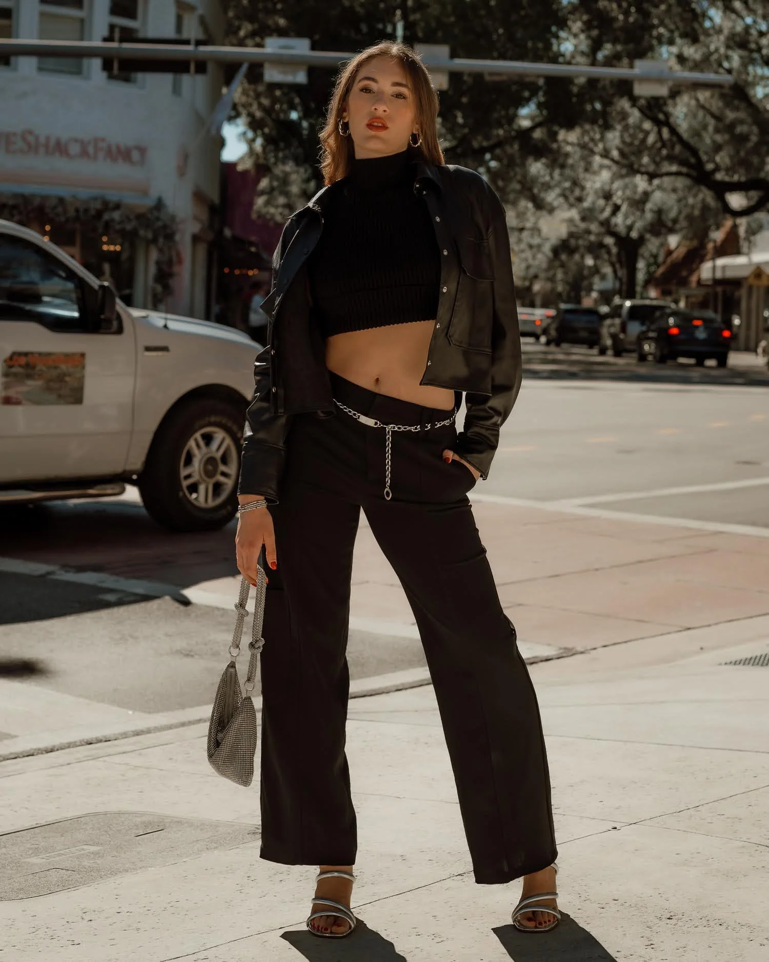A woman standing on a city sidewalk wearing a black cropped sweater, black pants with a chain belt, a black leather jacket, and silver high-heeled sandals, holding a small handbag.