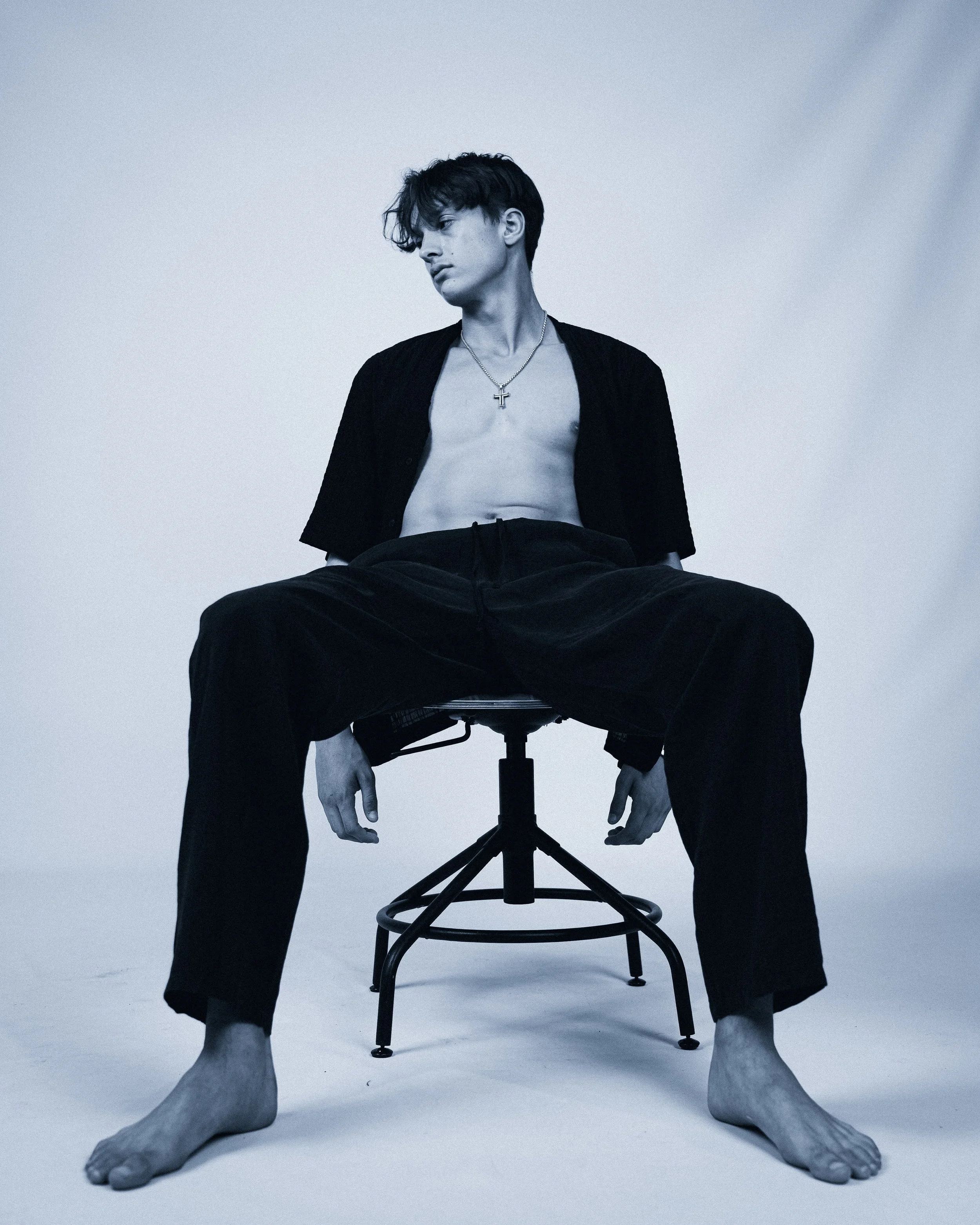 Black and white photo of a young man sitting on a chair, shirtless with an open shirt, wearing black pants and a cross necklace, with a contemplative expression.