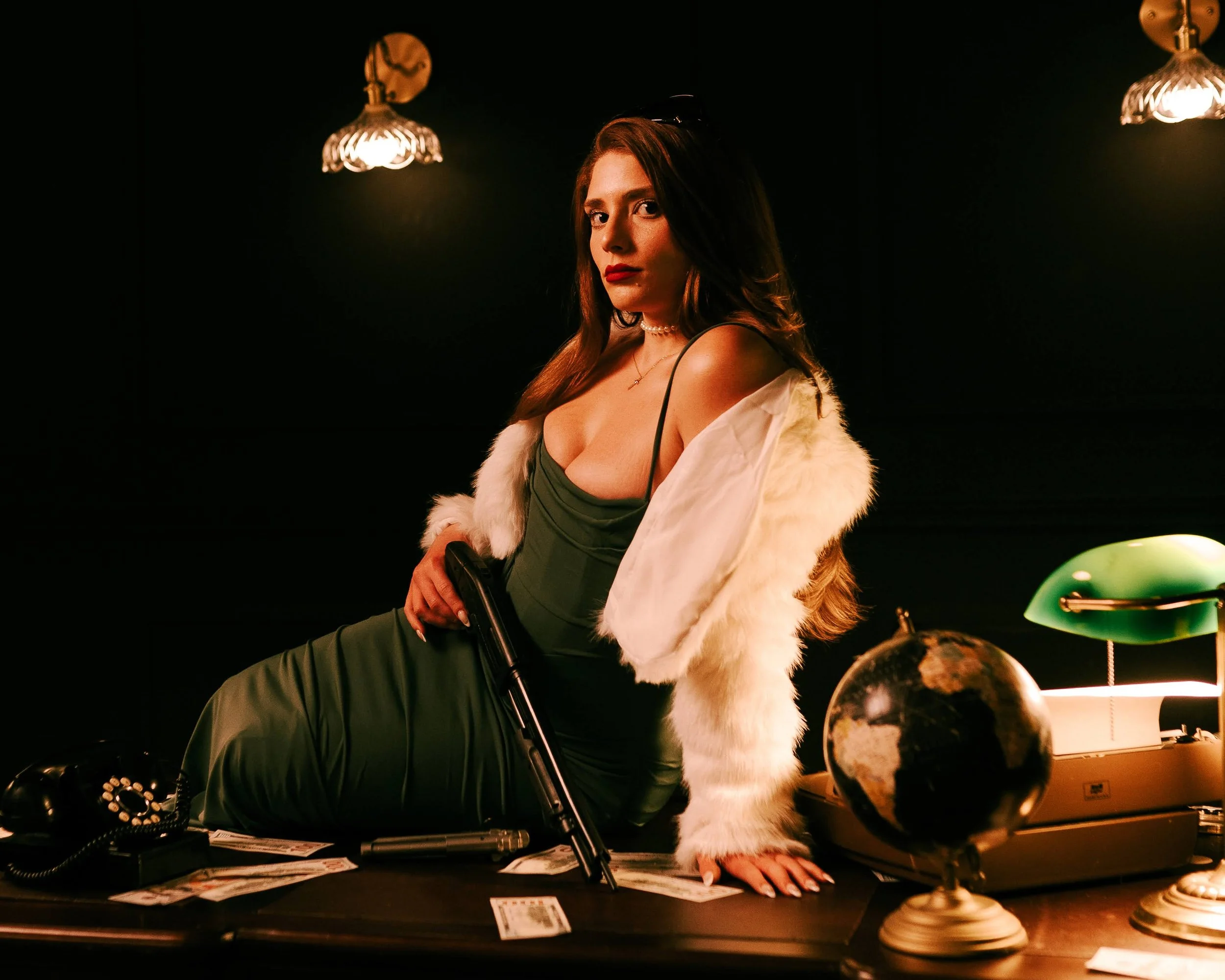 Woman in a green dress and white faux fur coat sitting on a desk in a dimly lit room with vintage office items and globe.