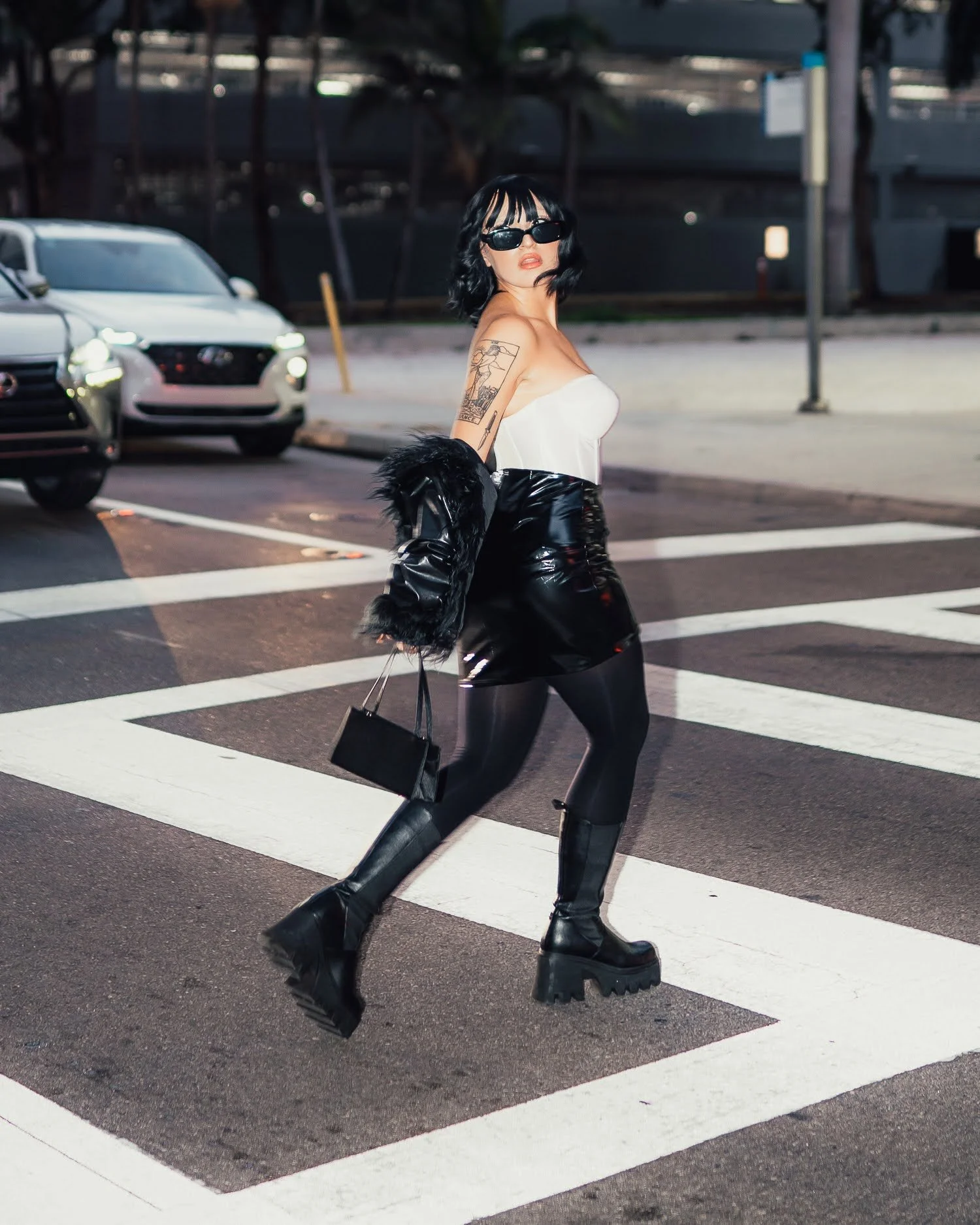 A woman with black hair, sunglasses, a white strapless top, black shiny skirt, black tights, and knee-high platform boots crossing a city street at night.