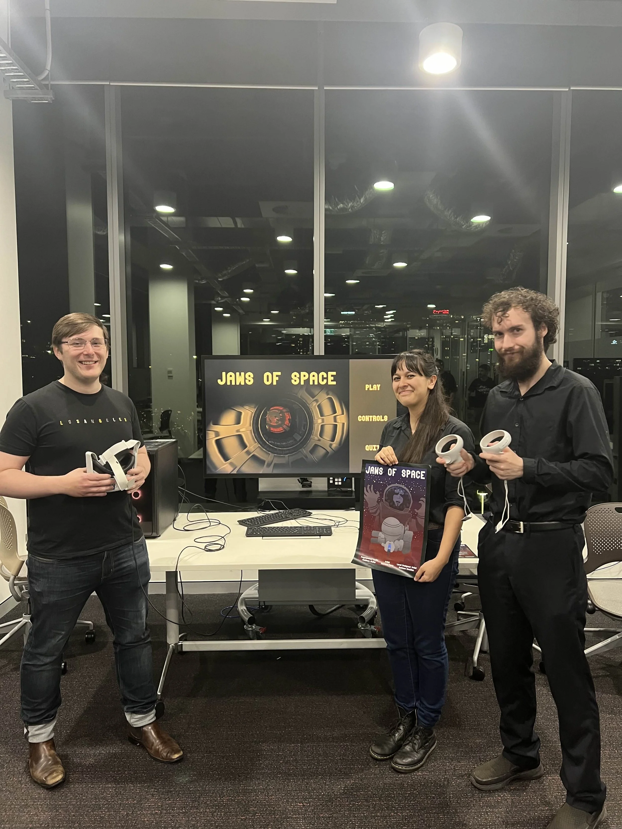 Three people standing in front of a TV screen displaying the game 'Jaws of Space' in a modern office. They are holding virtual reality controllers, and one woman holds a poster of the game.
