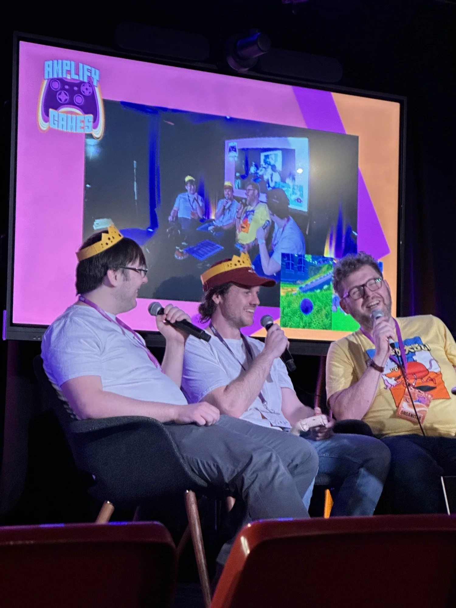 Three men sitting on stage at a panel event, each wearing a crown and holding a microphone, with a large screen behind them displaying an image of the panel from a different angle and the logo 'Amplify Games' in the top left corner.