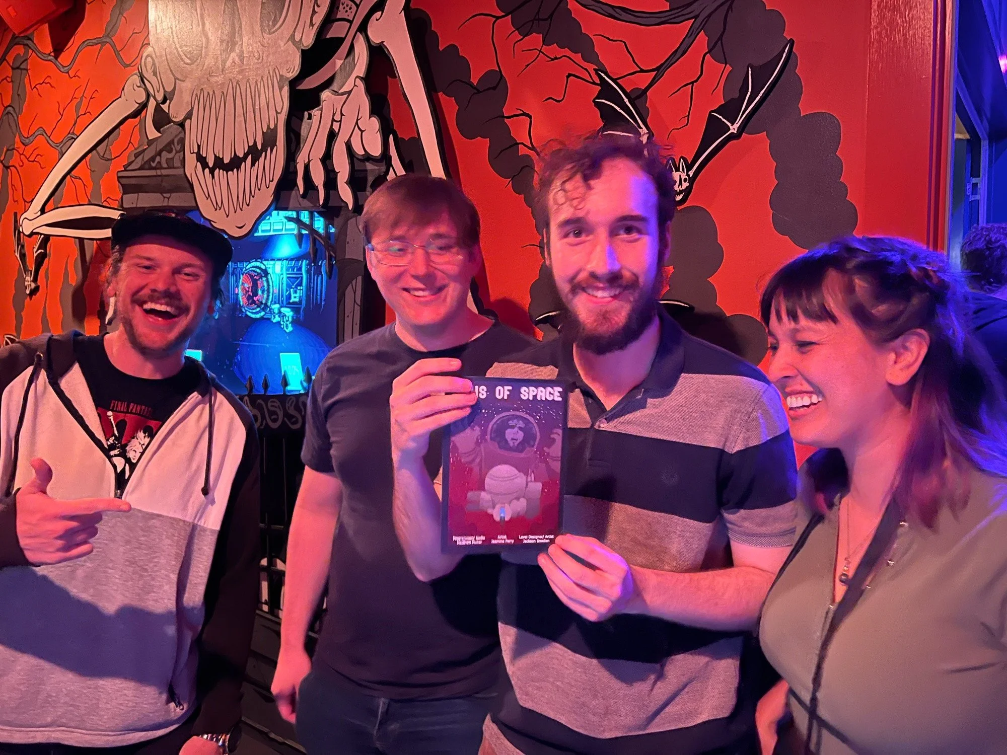 Four people smiling and standing together inside a themed arcade. One person is pointing at another who is holding a space-themed game. The background features a large skull mural with bats and a red and black color scheme.
