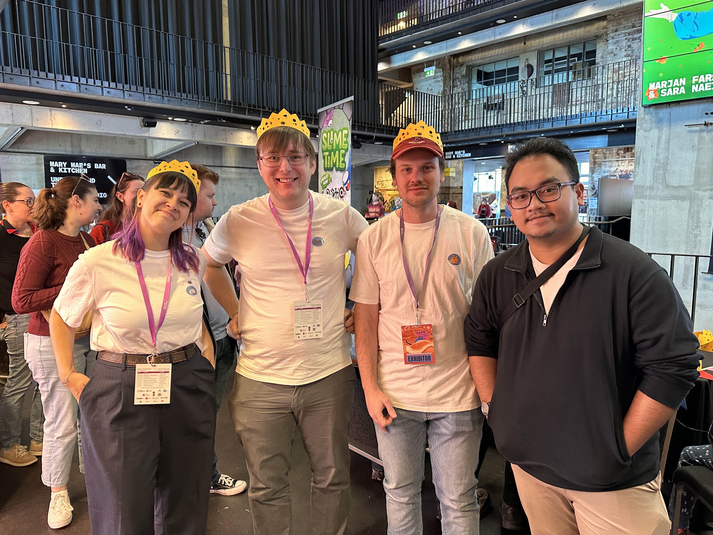 Four young adults standing together at an event, wearing yellow paper crowns and lanyards with badges, in a large indoor venue with several other people and some colorful signs in the background.