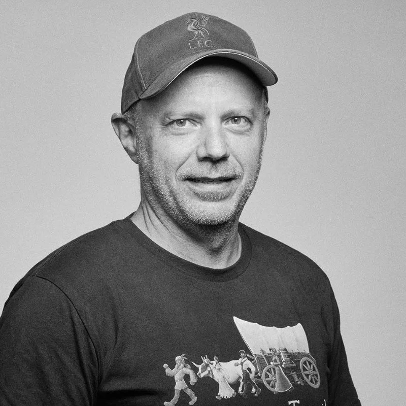 A middle-aged man wearing a baseball cap with a 'LFC' logo, a graphic t-shirt featuring a vintage circus scene, and looking directly at the camera with a neutral expression.
