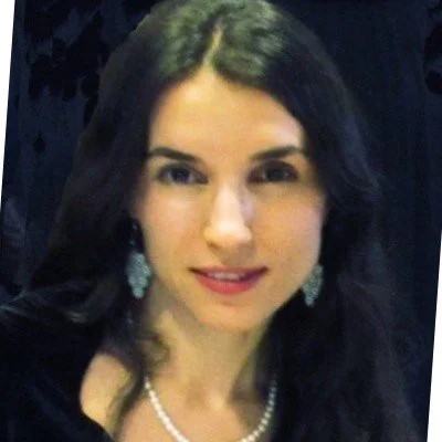 Portrait of a woman with dark hair wearing pearl jewelry against a dark background.