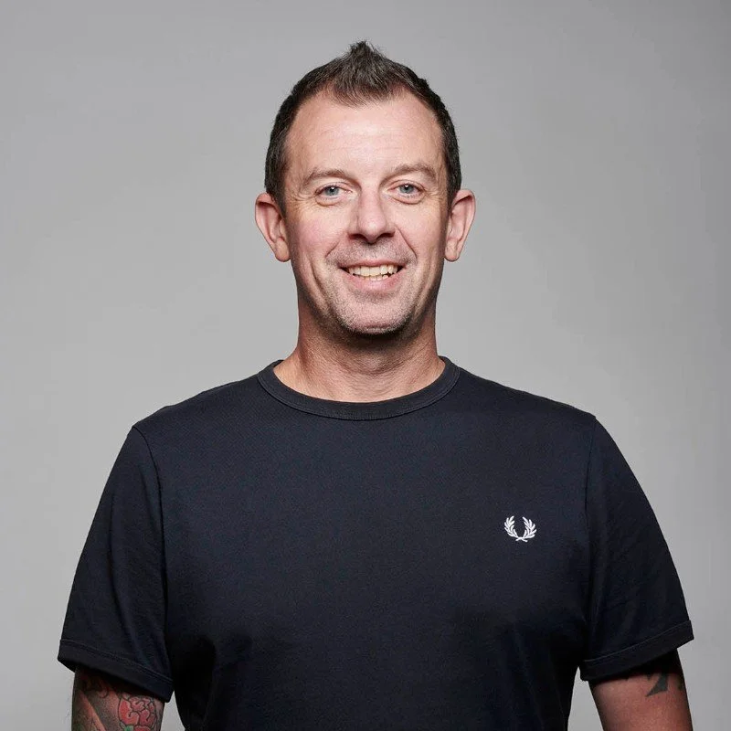 Portrait of a smiling man wearing a black t-shirt with a white logo, standing against a plain gray background.