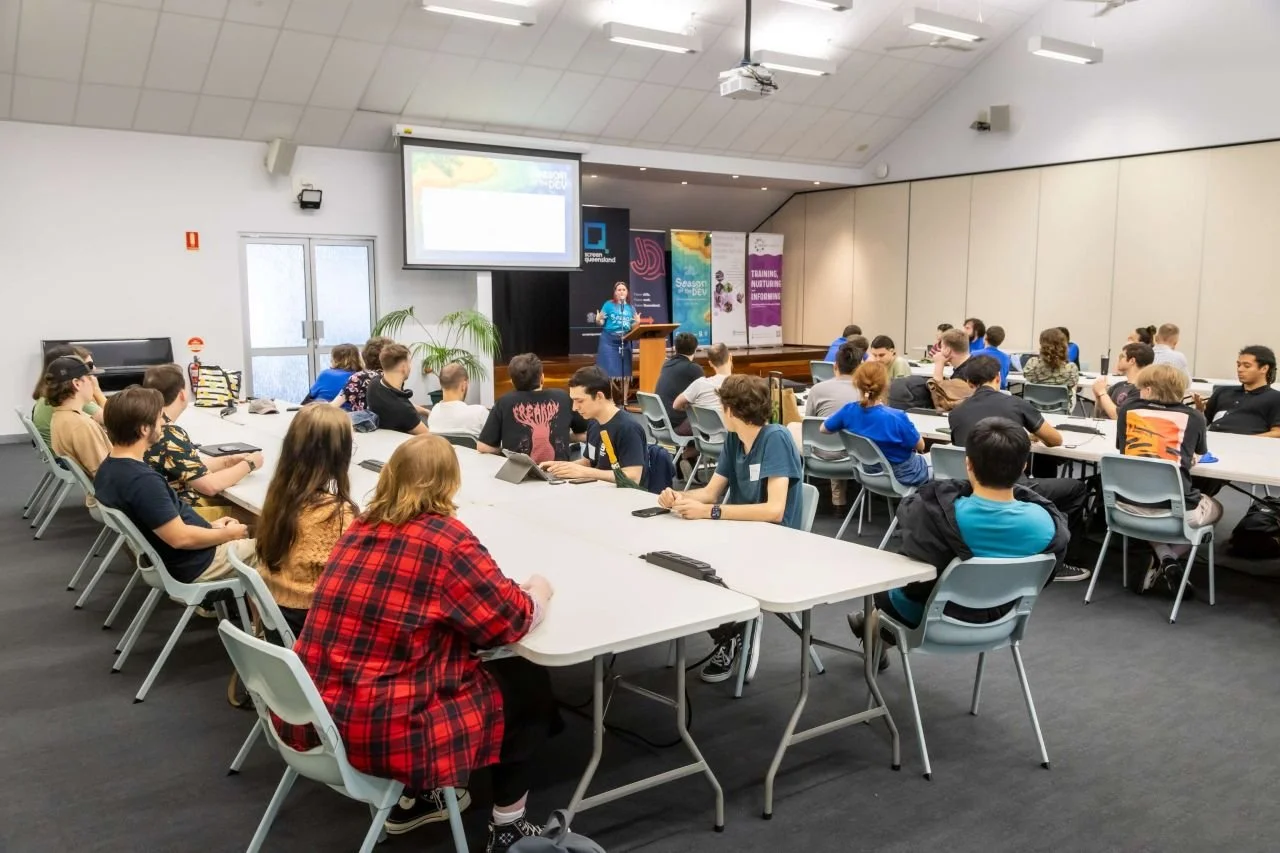 A classroom or conference room with students seated at long tables, listening to a presenter at a podium in front of a large screen displaying a presentation.