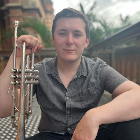 Young man sitting outdoors, holding a trumpet, with a faint smile, in a casual gray shirt, with plants and a fence in the background.