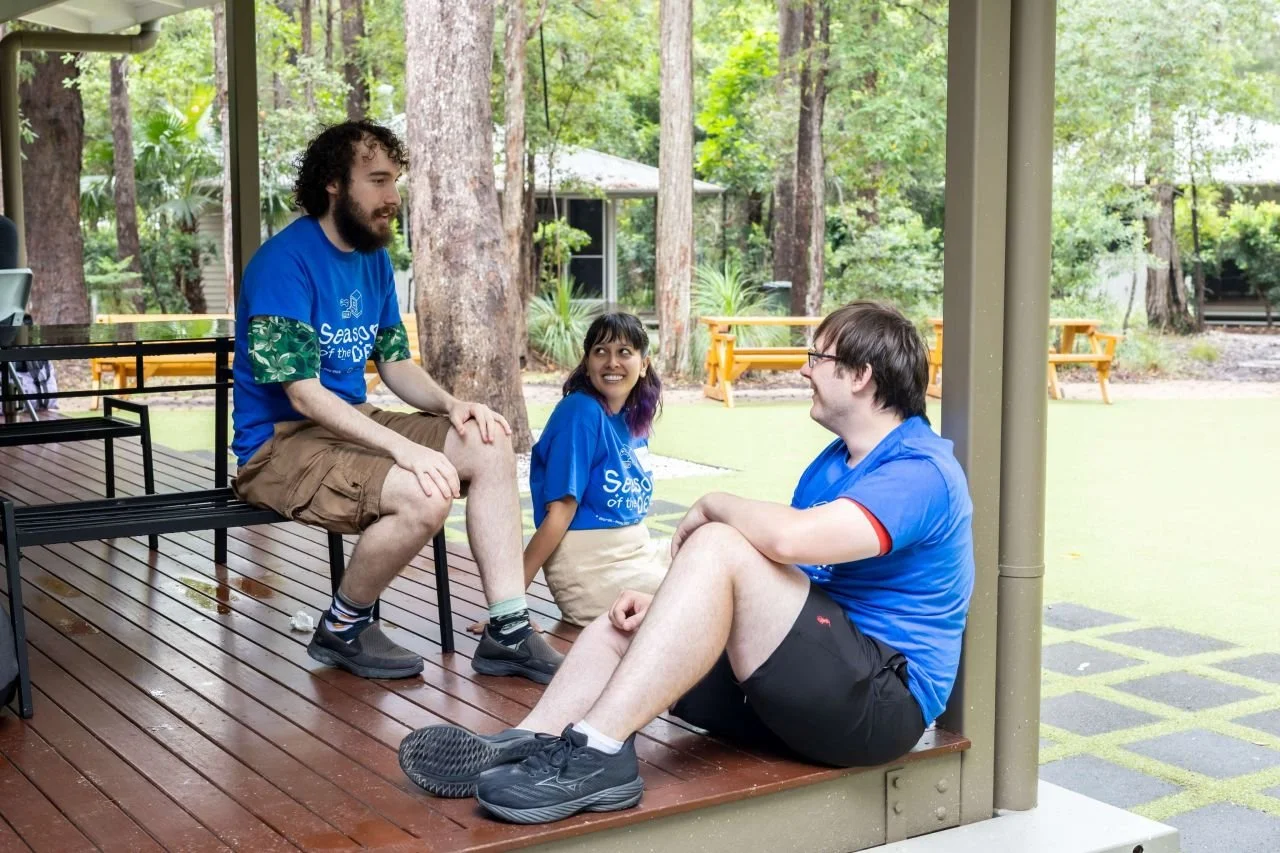 Three young people sitting on a wooden porch in a forested area, engaged in conversation and smiling, wearing blue shirts with white text, with trees and outdoor furniture in the background.