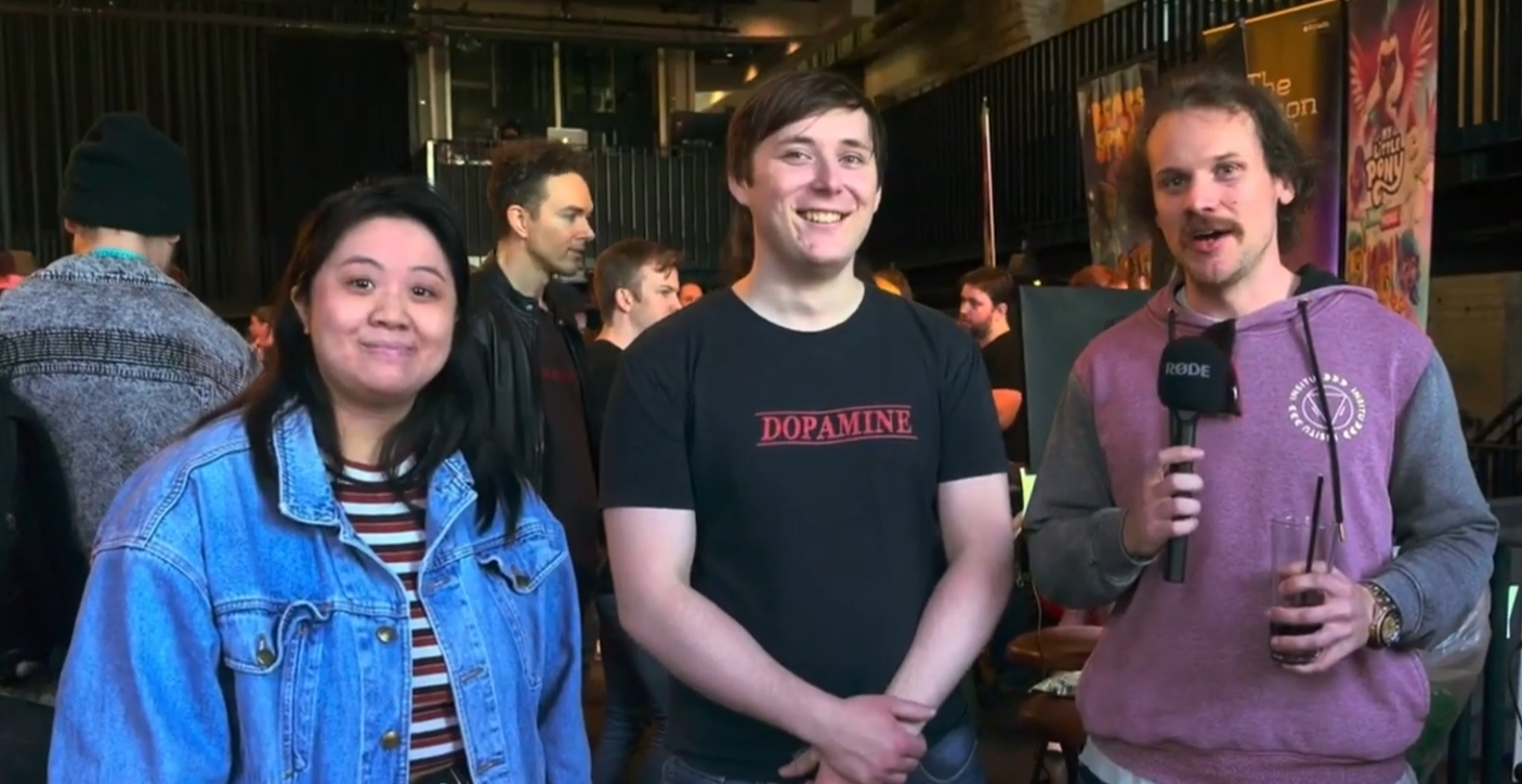 Three people standing together at an event, smiling at the camera, with a man holding a microphone on the right.