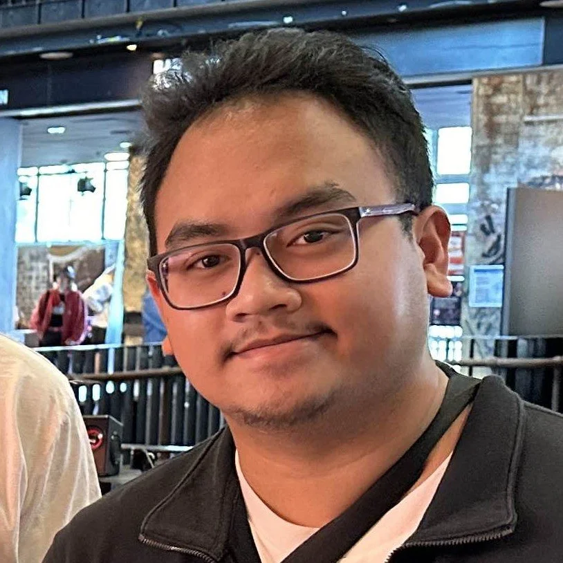 A young man with glasses and short dark hair smiling in a busy indoor setting with other people and large windows in the background.