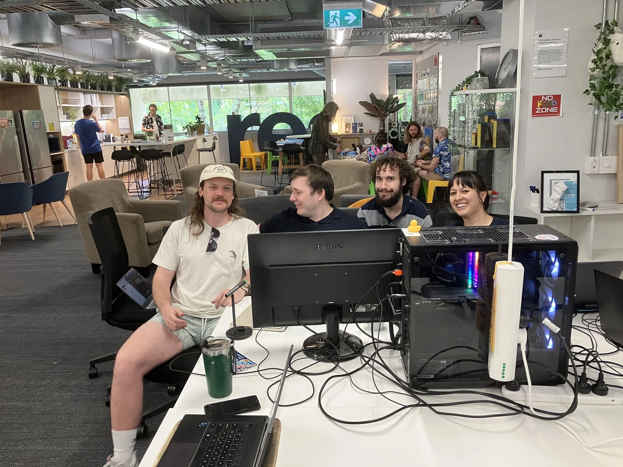 Four people seated at a table with computer equipment in a modern office space, with a kitchen area and other people in the background.