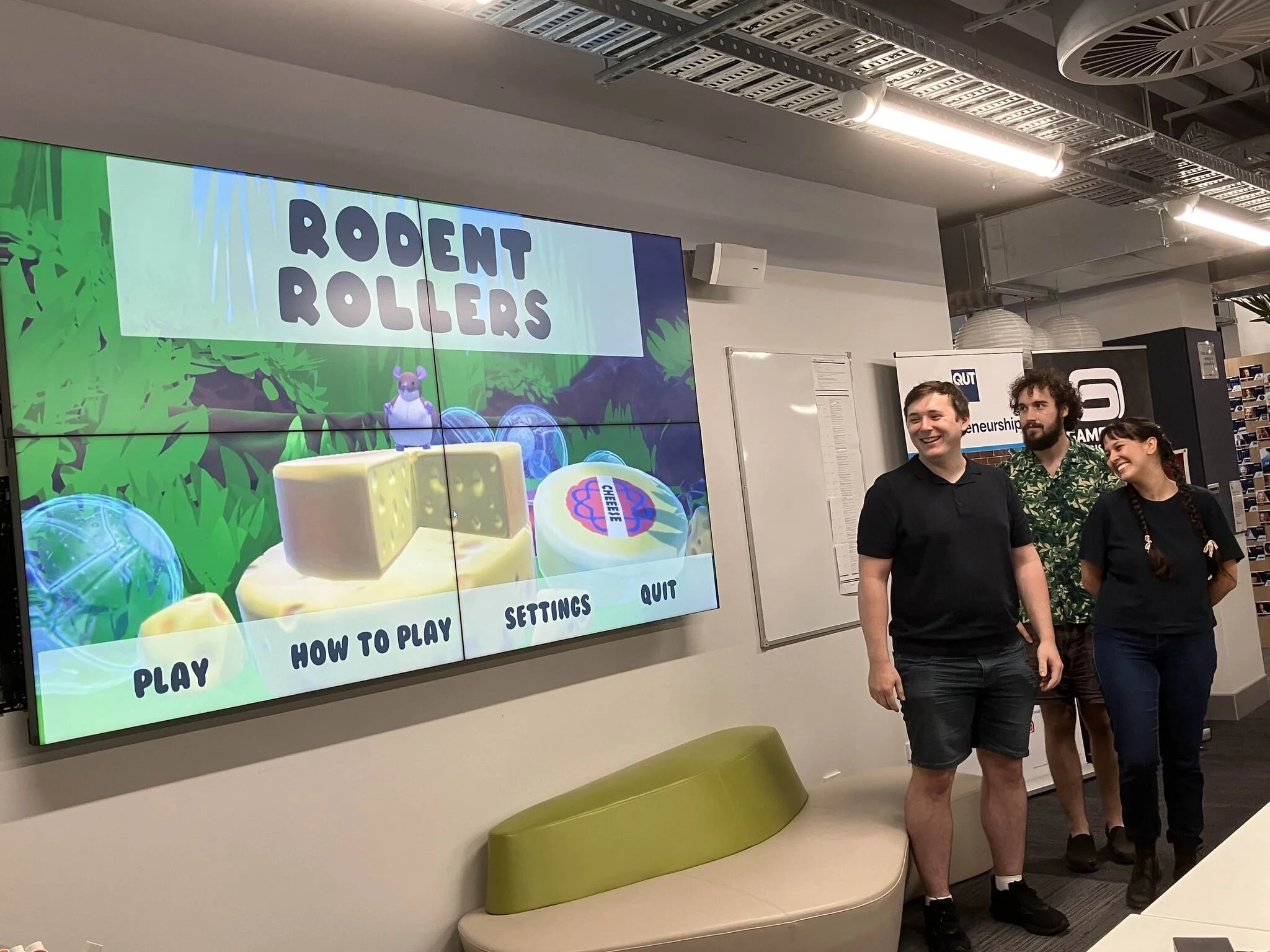 The image shows three people standing in front of a large screen displaying the game menu for 'Rodent Rollers.' The game screen features cartoon-style graphics of a mouse, cheese, and jungle foliage, with options for 'Play,' 'How to Play,' 'Settings,' and 'Quit.' The people are smiling