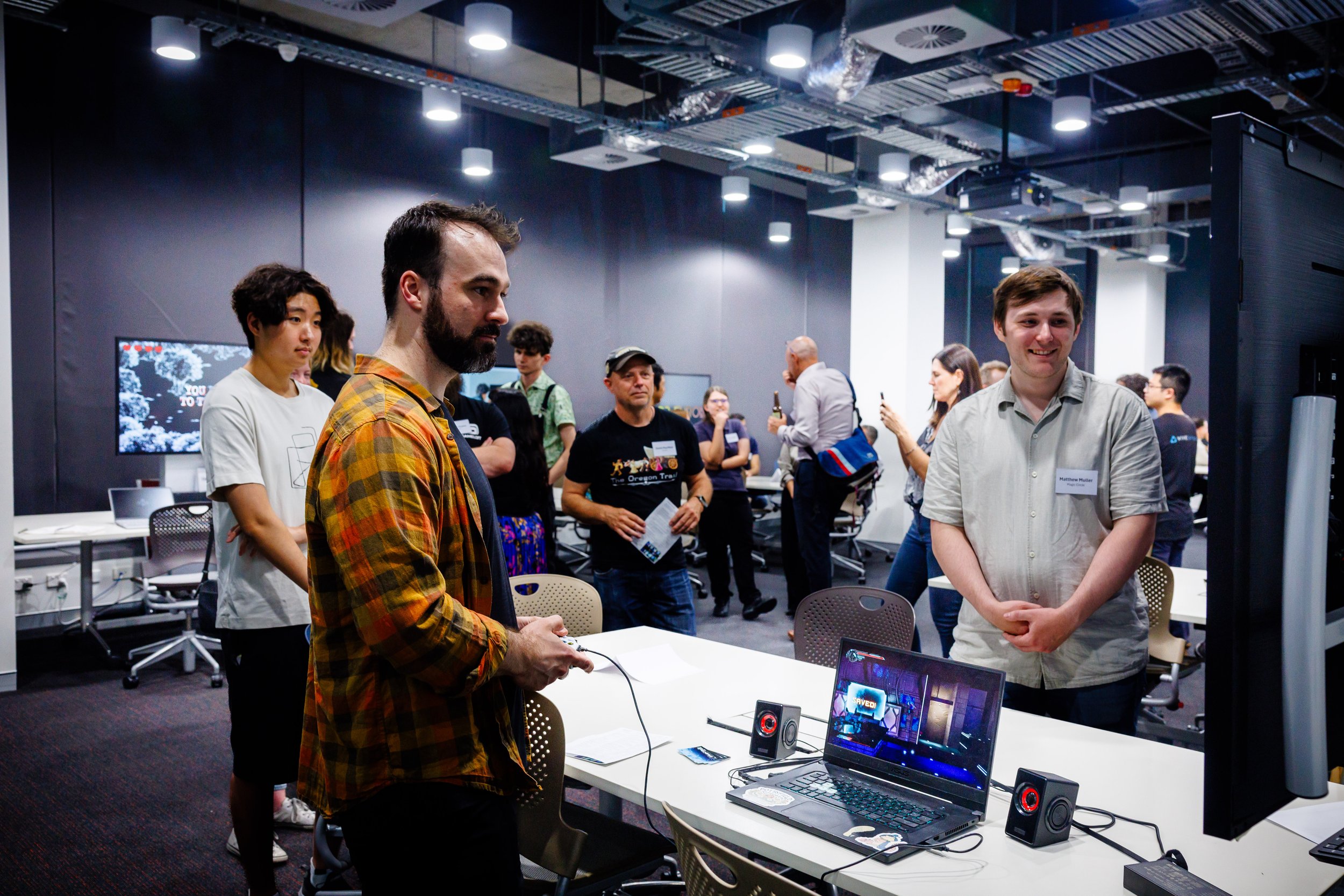 People gathered at a games showcase, with one person demonstrating a digital setup with a laptop and monitor on a table.