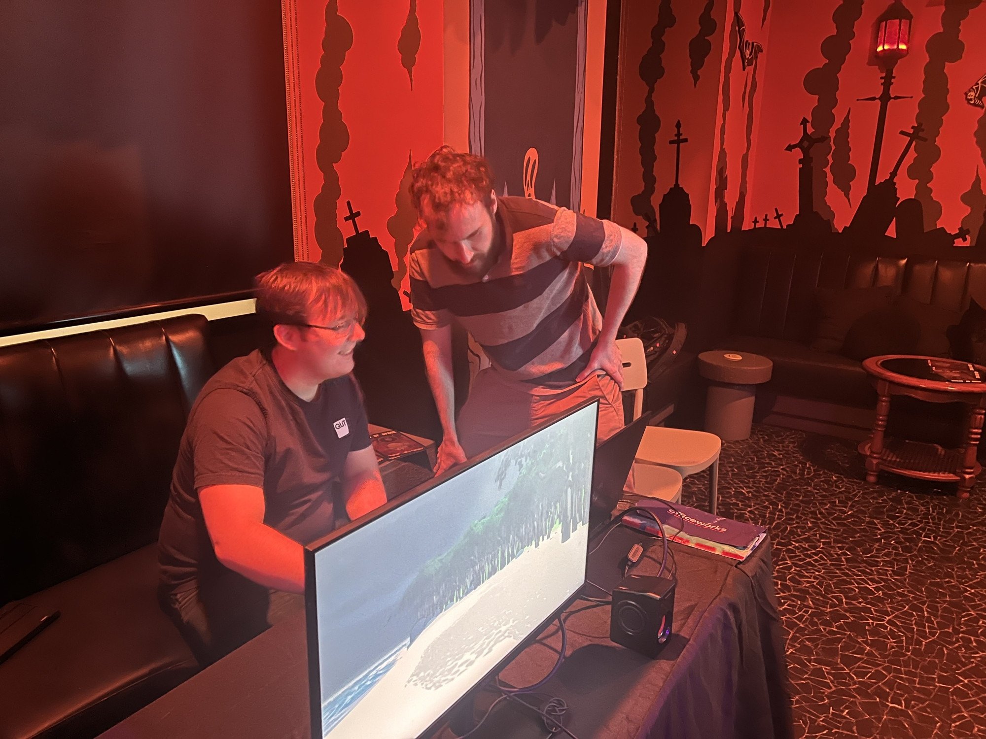 Two men are setting up a computer game in a dark, fiery themed room with silhouettes of crosses and smoke on the walls. One man is seated at a table with a computer monitor, and the other is standing, leaning over to look at the screen.