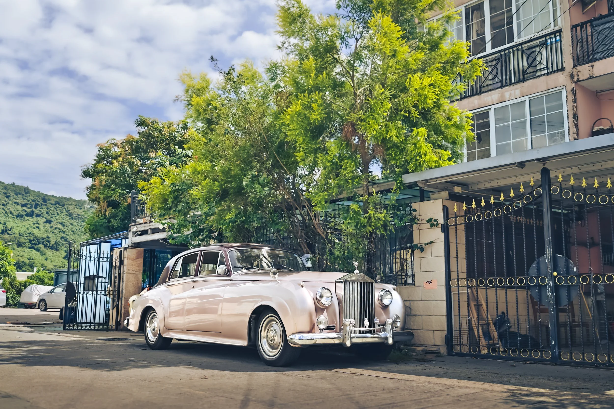 A vintage pink car parked on the street outside a building, with trees and hills in the background under a partly cloudy sky.