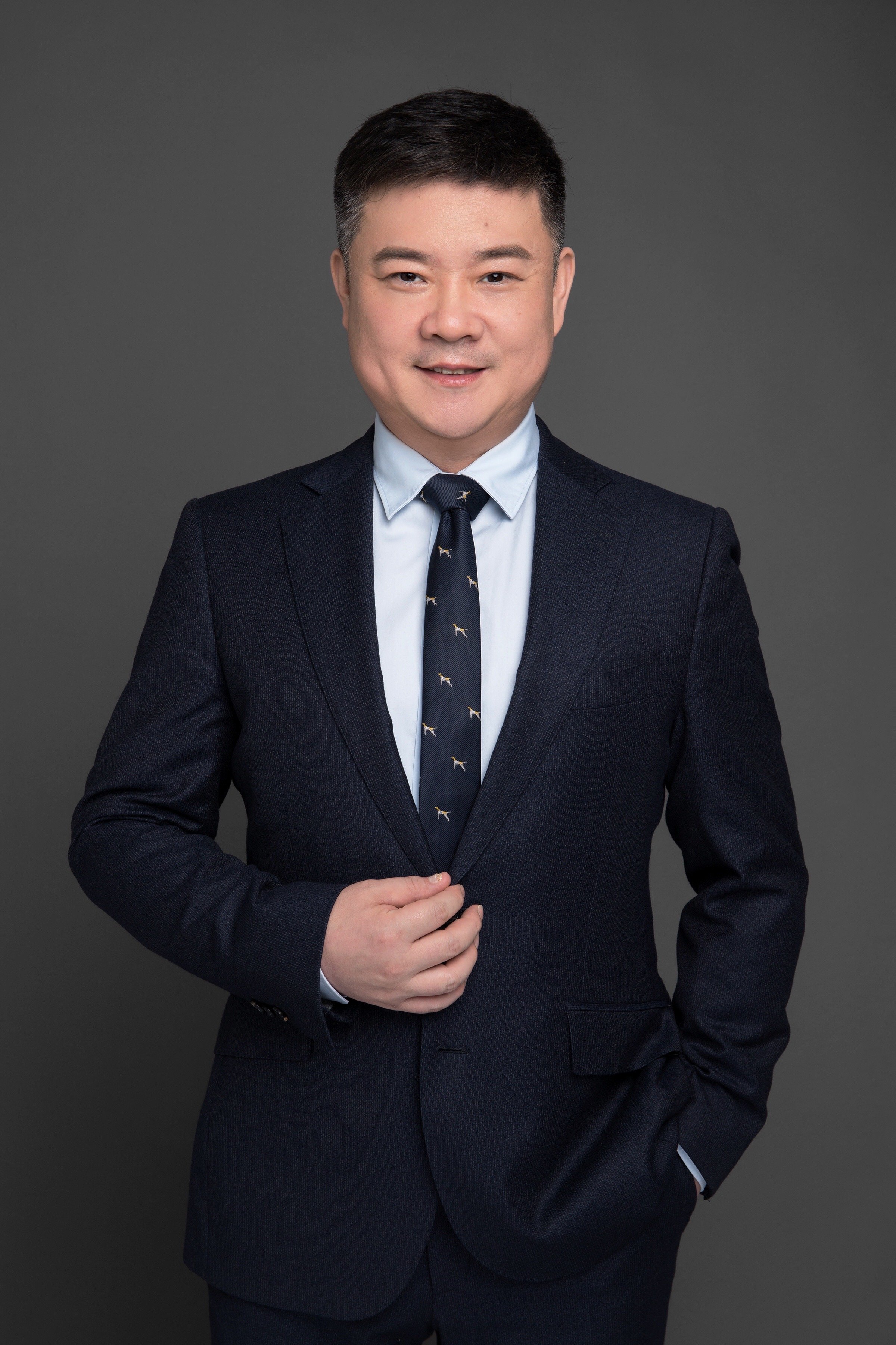 A man in a dark suit with a light blue shirt and a tie featuring small dog patterns, posing against a gray background.