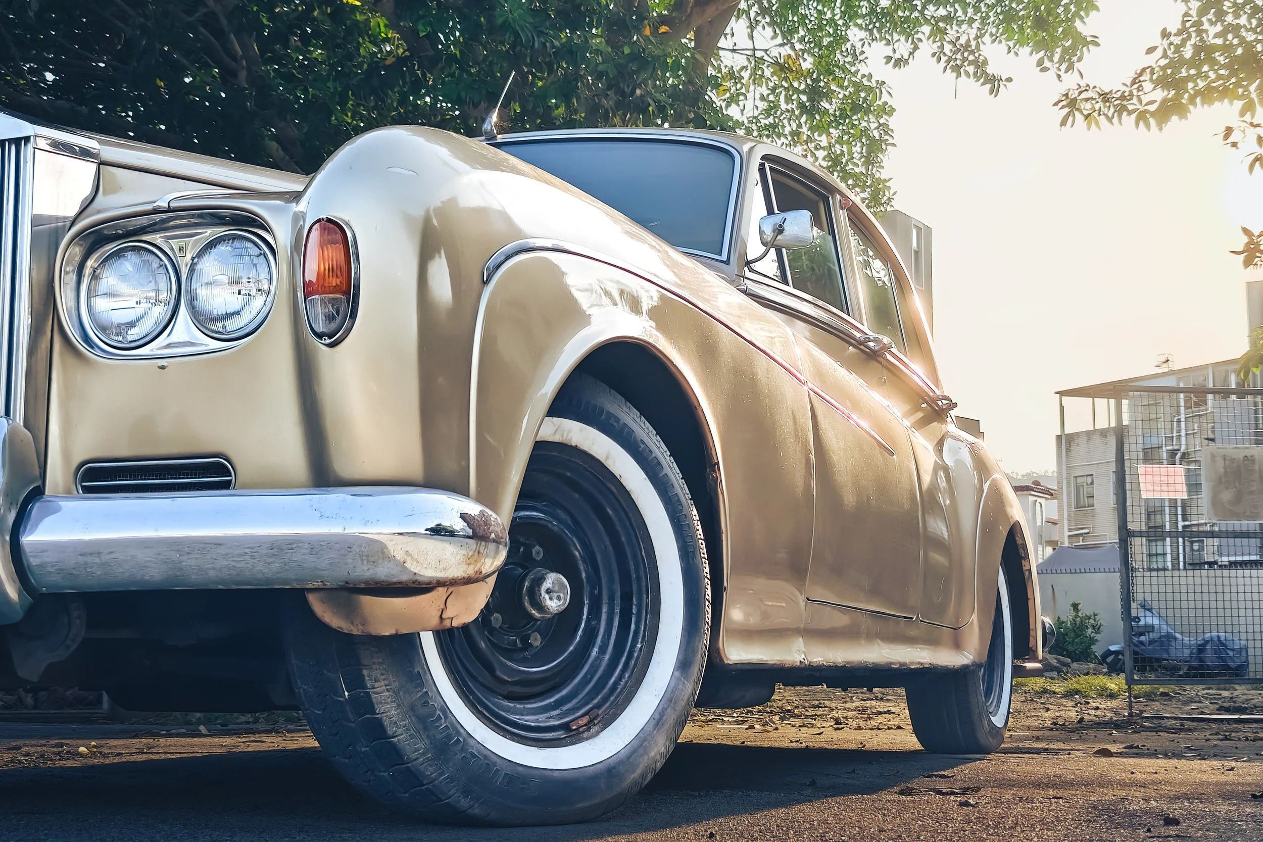 A vintage beige car with black and white tires parked outdoors near some trees and buildings, captured in sunlight.