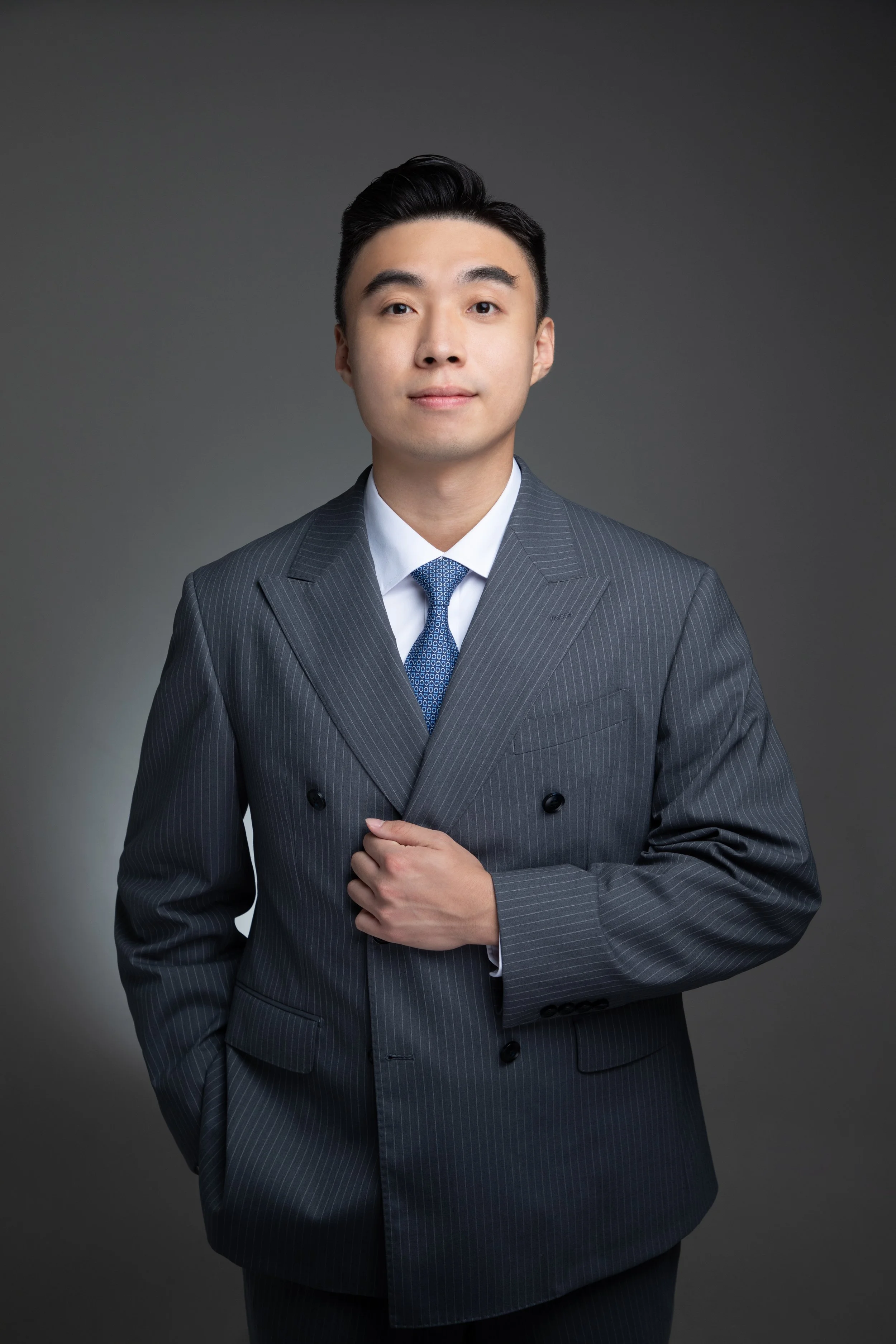 A man in a gray suit with arms crossed, wearing a blue tie and watch, standing against a white background.