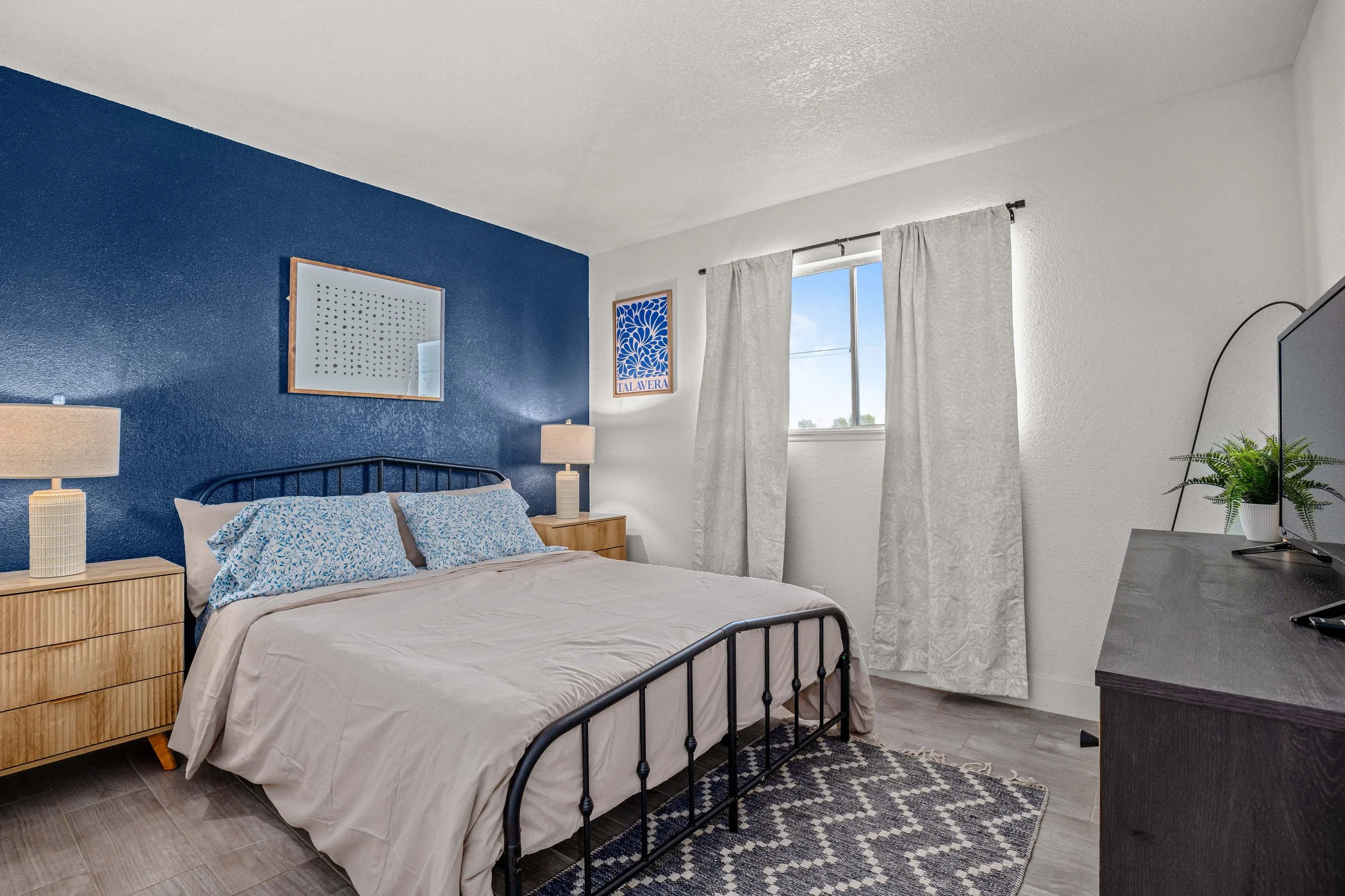 A bedroom with a dark blue accent wall, a bed with pillows and beige bedding, wooden nightstands with lamps, white curtains over a window, a dresser with a television and a potted plant.
