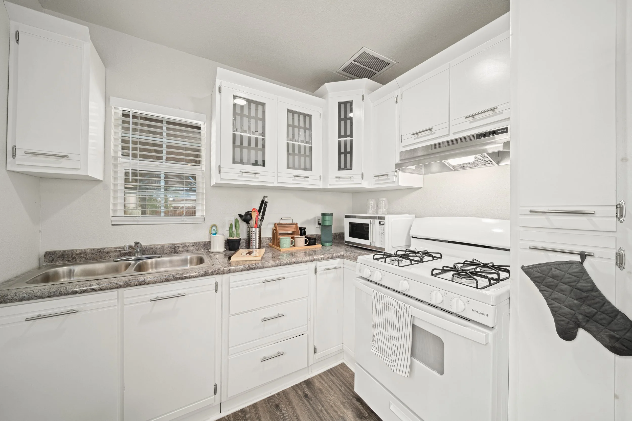 White kitchen with granite countertops, double sink, white cabinets, microwave, gas stove, oven, and a black oven mitt hanging from the oven handle.