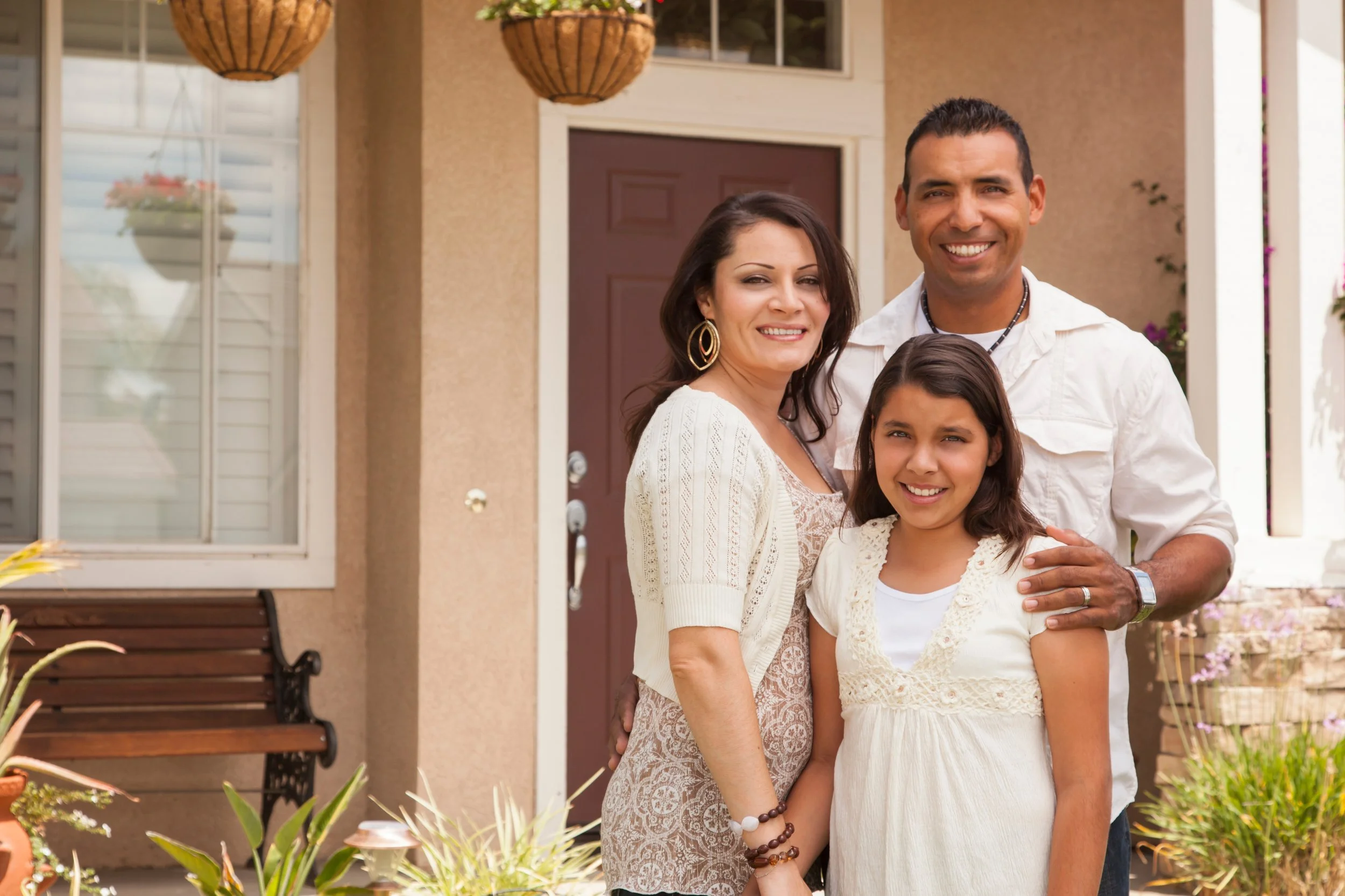 A smiling family of three standing outside their house, with a woman and man embracing a young girl. They are dressed casually and look happy.