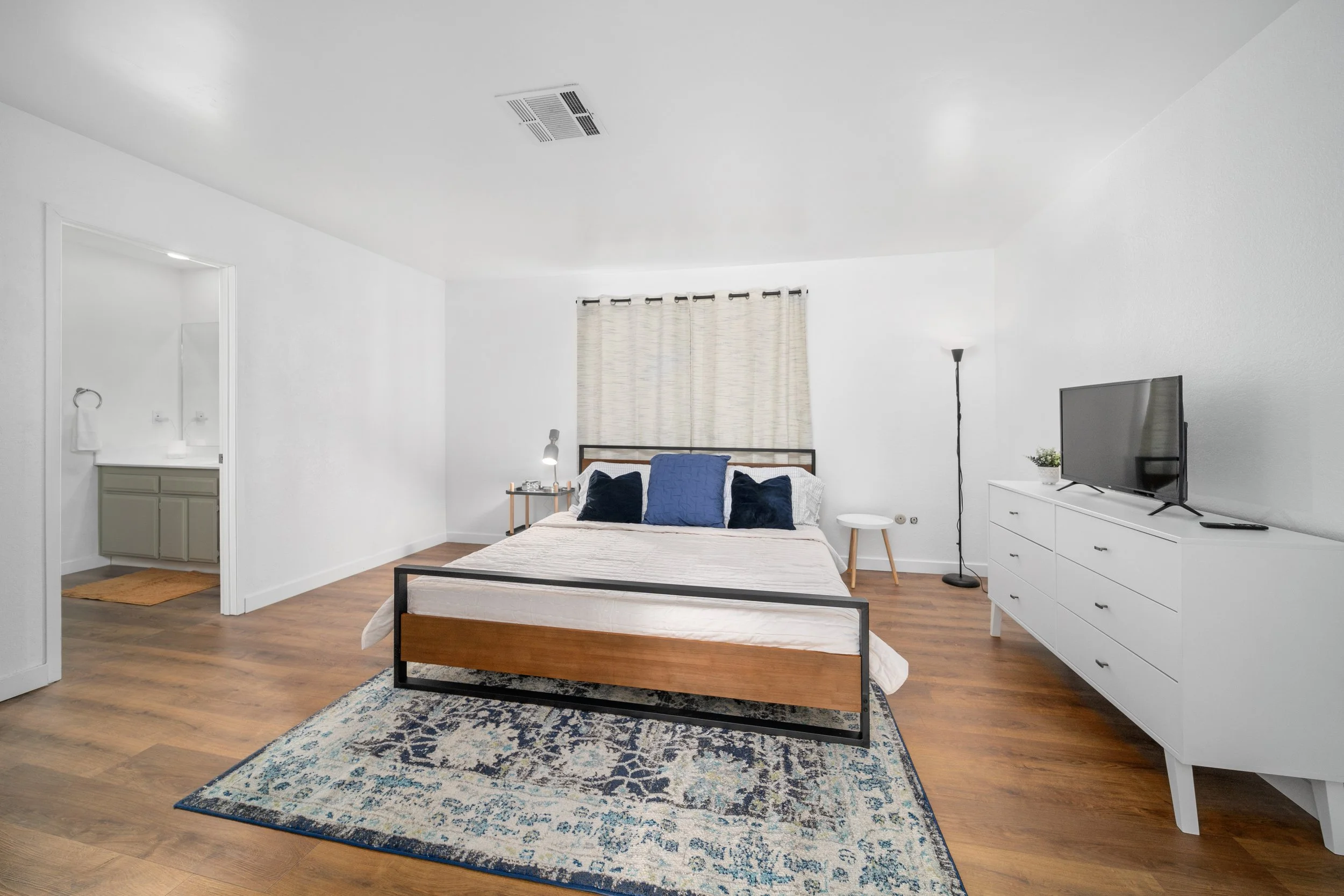 A modern bedroom with white walls, wooden flooring, a large bed with navy and white pillows, a white dresser with a TV, a tall floor lamp, a side table, and a small bathroom visible through an open door.