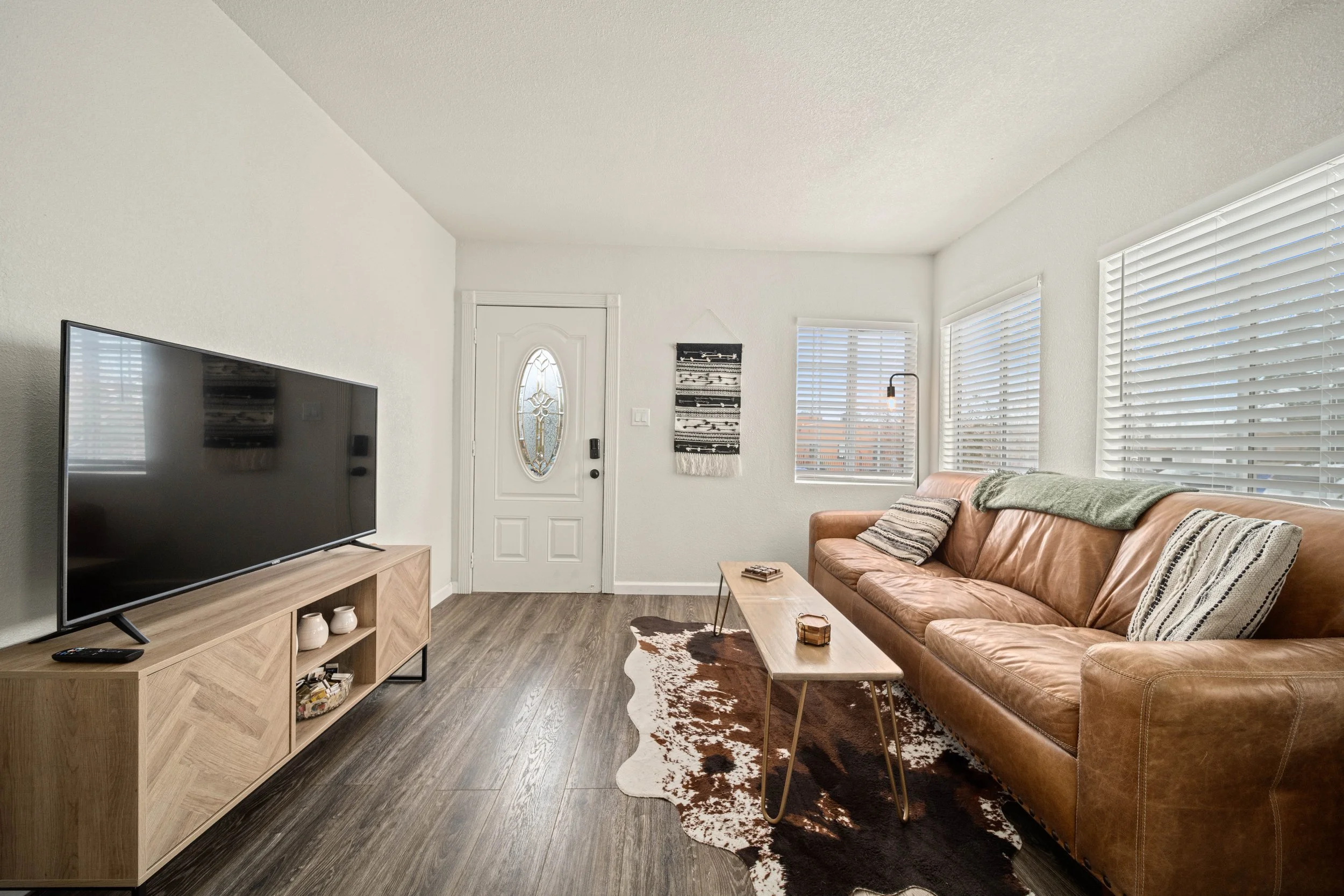 Living room with a brown leather sofa, a flat-screen TV on a wooden stand, a small wooden coffee table, and standing lamps near windows with white blinds. The room has light-colored walls, hardwood flooring, and a cowhide rug.