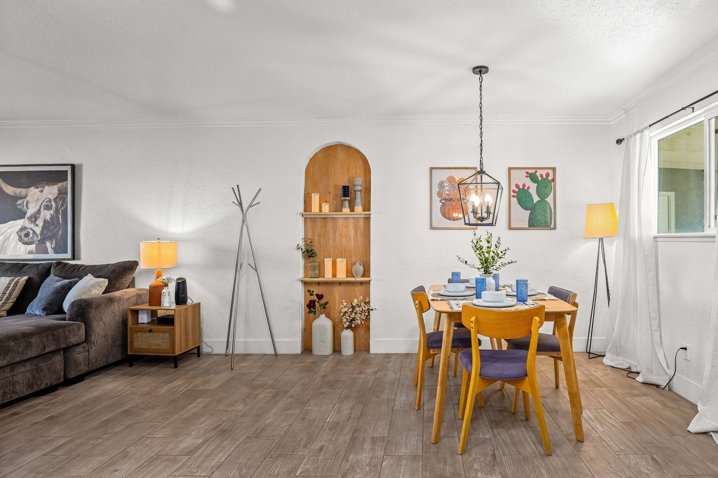 A living and dining area with a gray sofa, dining table set with plates and glasses, wall art, and various decorative items and lighting.