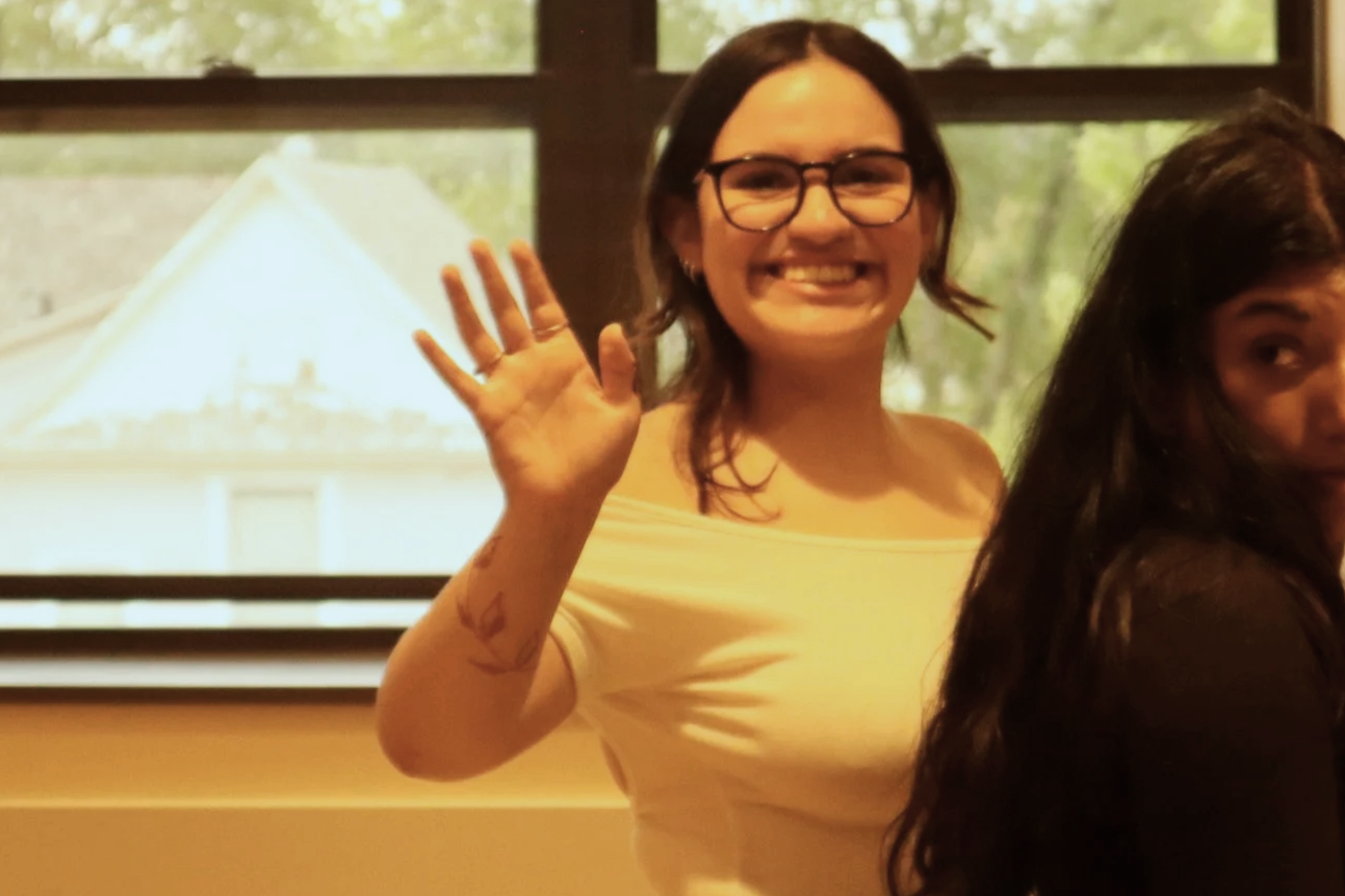 A woman with glasses and shoulder-length dark hair smiling and waving, sitting indoors next to another woman with long dark hair. Behind them is a window with a view of a house and trees.