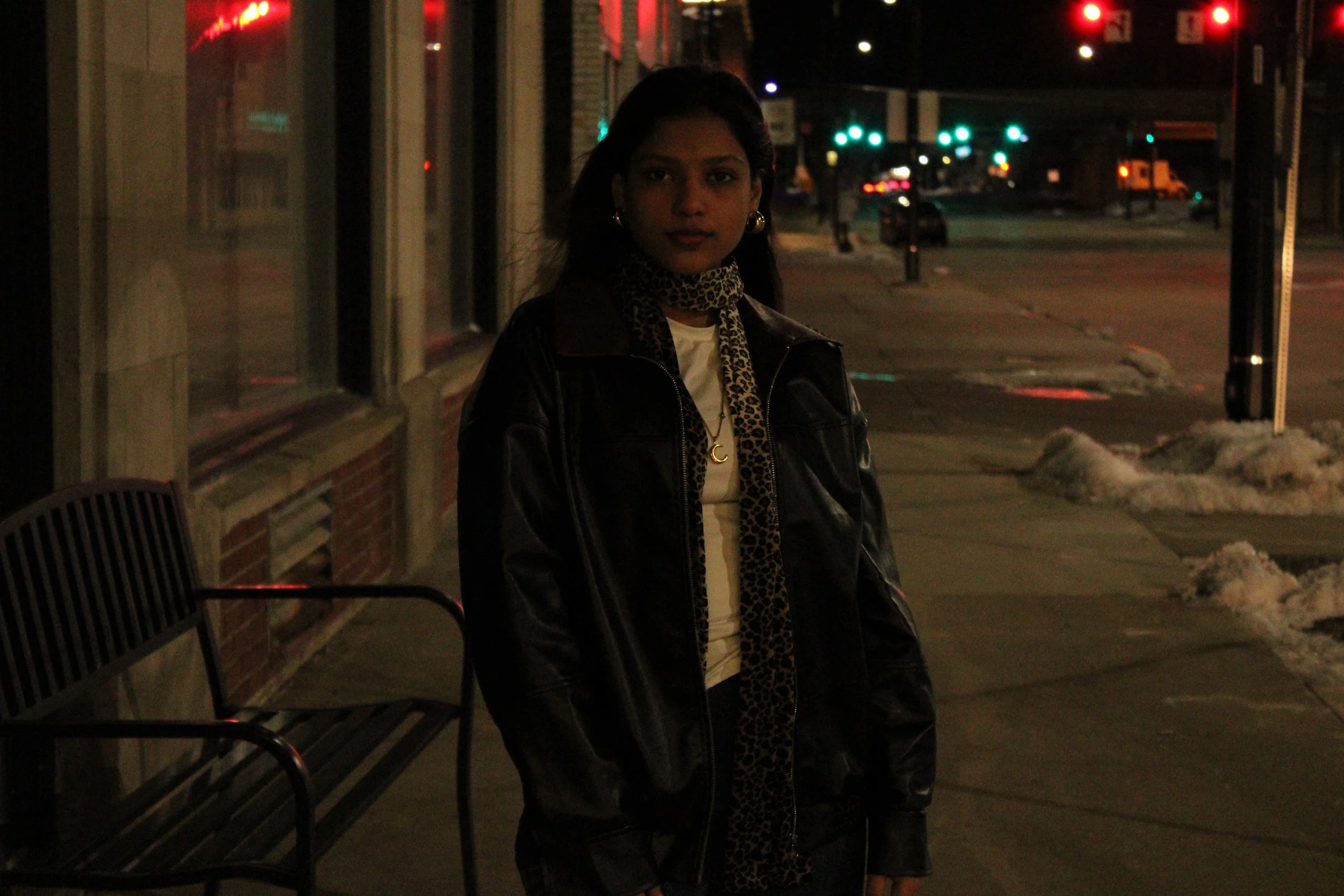 A young woman standing on a sidewalk at night, wearing a black leather jacket and leopard print scarf, with snow on the ground and city lights in the background.